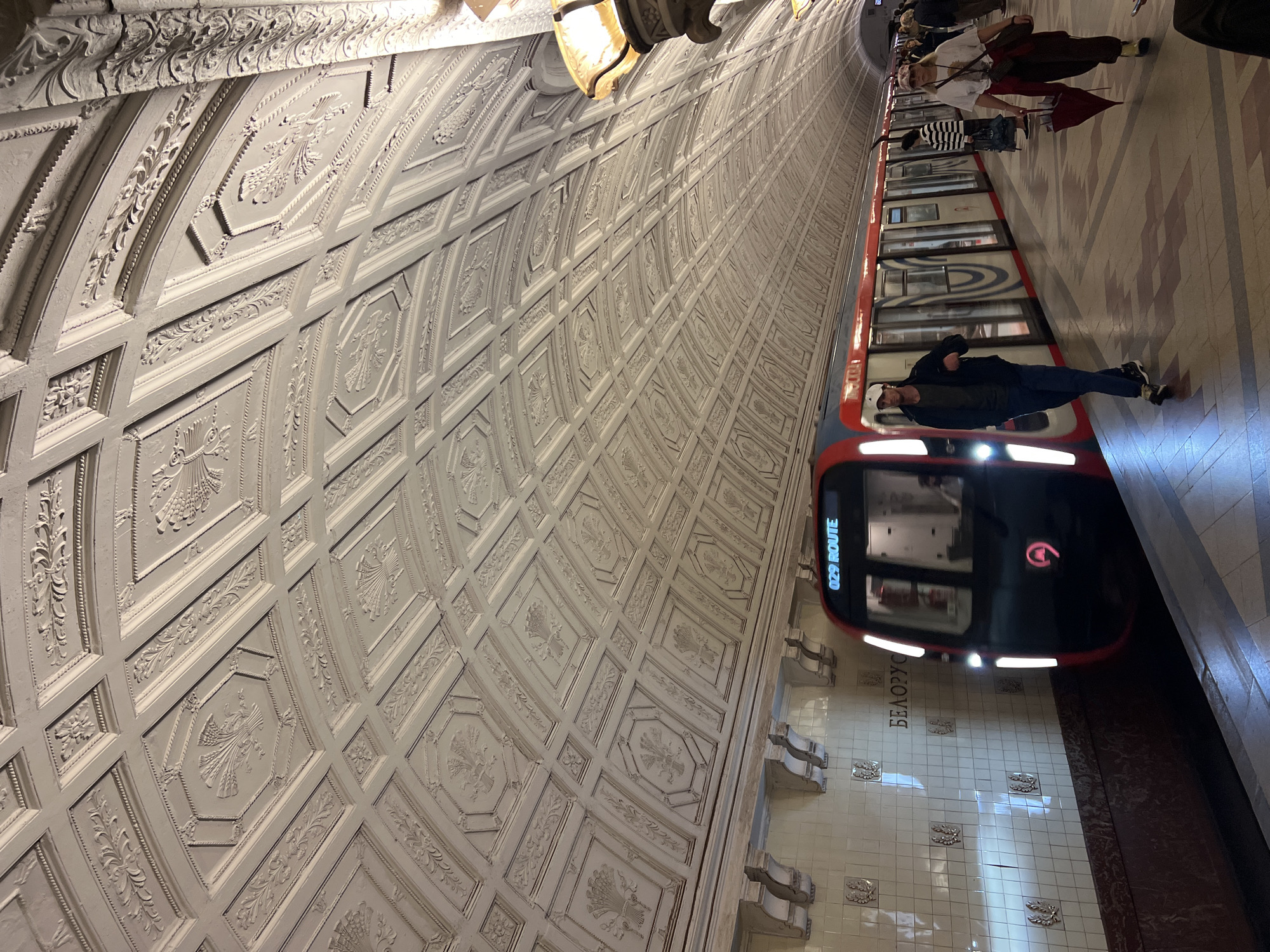 This is a photograph of a Moscow Metro station platform. The station is adorned with ornate, white coffered ceilings featuring relief carvings of floral and geometric patterns. The tiled walls of the station are white, with some decorative elements and the station name "БЕЛОРУССКАЯ" (Belorusskaya) visible. A modern red and white train with the designation "029 ROUTE" displayed on its front is arriving at or departing from the platform. Several people are present on the platform, some walking, others standing. One man in a dark jacket and a white cap is walking towards the camera, while a woman in a white t-shirt and dark pants, holding a red umbrella, is walking away from the camera. The platform floor is tiled with a pattern of red and beige squares. The lighting in the station appears to be a mix of artificial light from fixtures and ambient light from the train.
