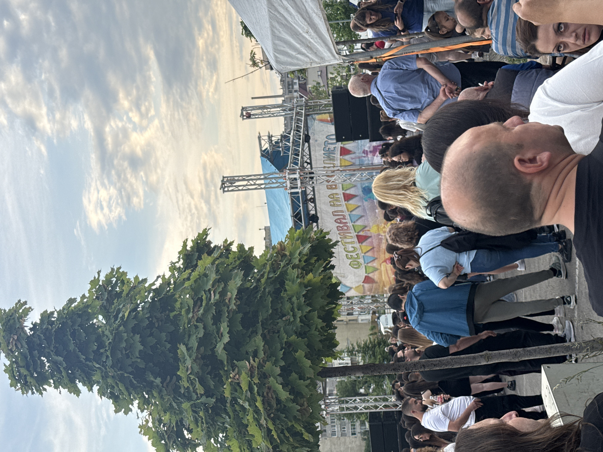 A crowd of people gathers in Shumen, Bulgaria, under a partly cloudy sky, facing a stage setup for the "Festival Na Vhusto". The scene includes a large tree on the left side and metal scaffolding for the stage. Individuals are seen standing, looking toward the stage, with some faces visible in the foreground. Buildings are visible in the background, and the overall setting suggests an outdoor public event.