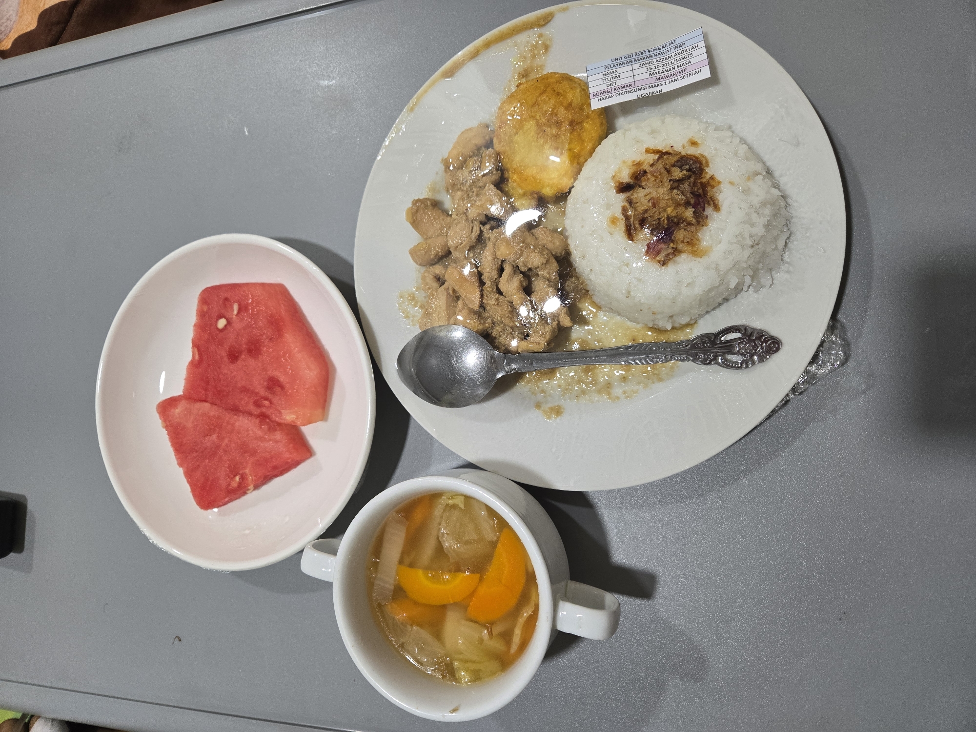 A meal is presented on a gray surface, likely a tray table. The meal consists of a white plate with a serving of rice, a portion of what appears to be stir-fried chicken, and a fried egg. The plate is partially covered with plastic wrap. A spoon rests on the plate. To the left of the main plate is a white cup containing a clear broth with visible pieces of orange carrot and white vegetables, likely soup. Above these is a small white bowl with two triangular slices of watermelon. A label with text, partially visible and appearing to be in Indonesian, is attached to the side of the main plate. The text on the label indicates it is from the "UNIT GIZI RUMAH SAKIT SUNGAILIAT" which translates to "Nutrition Unit of Sungailiat Hospital". The presence of the hospital name and location suggests the meal was served in Sungailiat, Indonesia. The overall environment is clean and appears to be a hospital setting, possibly a patient's meal.