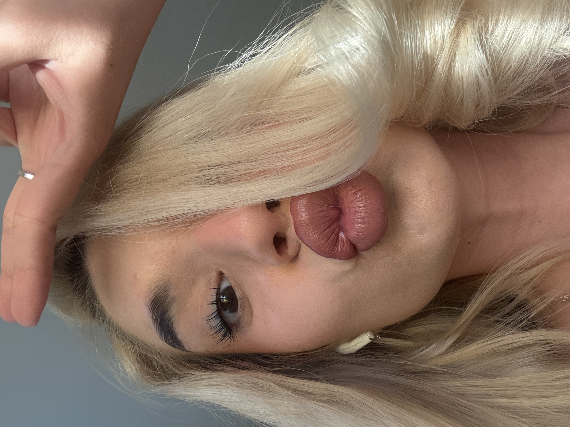 A close-up portrait of a woman with blond hair. She is puckering her lips in a kissing gesture. Her eye is brown, and she has dark, well-defined eyebrows. She wears makeup, including eye shadow and lip gloss. Part of her hand is visible at the top left corner, with a ring on her finger. A small earring is seen near her ear. The background is a light grey. No information about the city of Sofia, Bulgaria is available in the image.