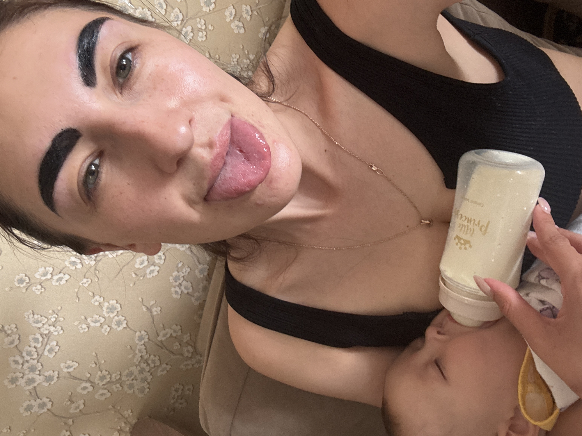 A woman is lying on a floral patterned sofa, holding a baby bottle up to a baby's mouth. The bottle is labelled "Little Princey". The woman is wearing a black tank top and a gold necklace. She has thick, dark eyebrows and is sticking her tongue out. The baby is wearing a bib. The environment appears to be indoors. There is no information available to determine the location in Sochi, Russia.