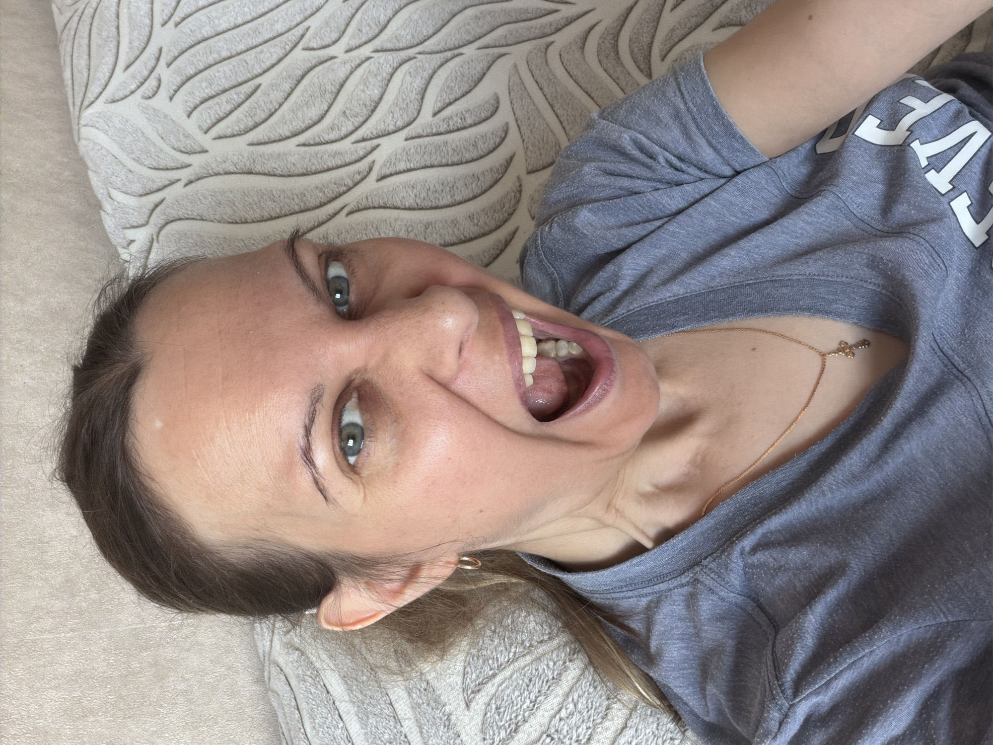 A woman is lying on a beige patterned pillow or cushion, angled toward the upper left corner of the frame. She is wearing a grey short-sleeved shirt with white lettering that is partially visible. The woman has fair skin, light-colored eyes, and brown hair pulled back from her face. Her mouth is open wide, revealing her teeth. She wears a thin gold necklace with a pendant that rests on her collarbone. There is an earring in her left ear. There is no indication of this image being taken in Izhevsk, Russia.