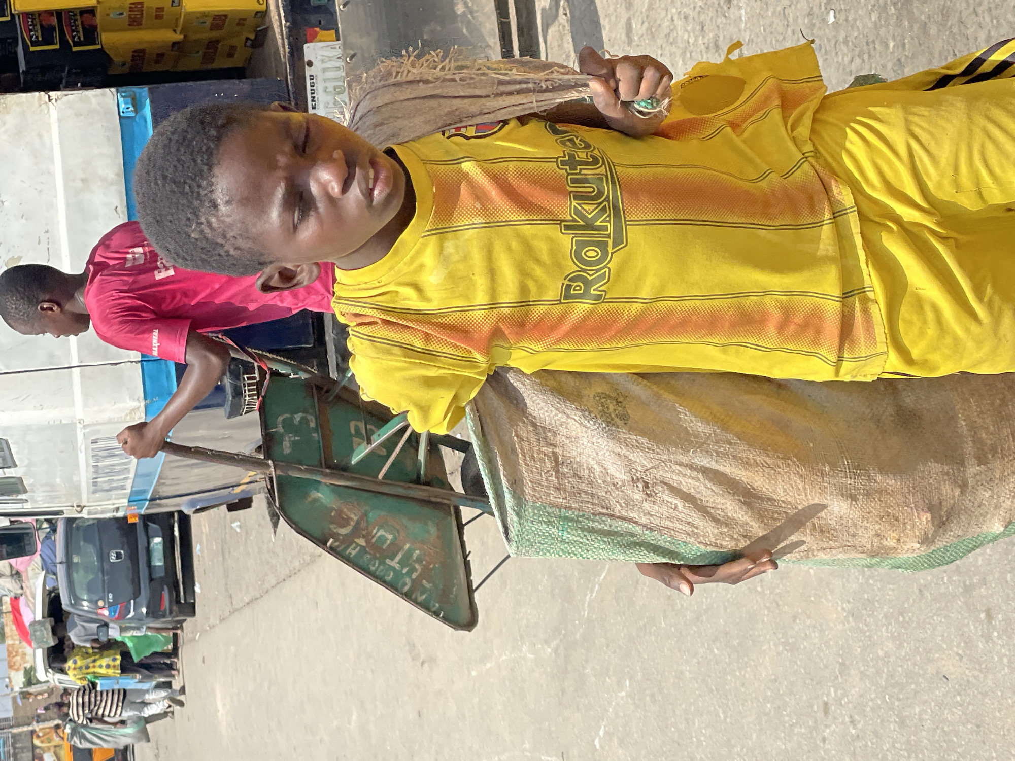 The image captures a young Black boy in Fegge, Nigeria, appearing to be in a crowded outdoor market or transportation hub. He is wearing a vibrant yellow t-shirt with orange stripes and a "Rakuten" logo, and also a pair of yellow shorts. His eyes are closed, suggesting he might be resting or tired, and he is holding a bundle of what appears to be dried straw or hay in his right hand.
Behind him, another young Black person in a bright pink t-shirt is visible. The boy is standing near a wheeled cart, which is partially obscured by a large, rough burlap sack, also containing what looks like dry plant material. In the background, there are glimpses of vehicles, including a dark-colored car, and what seem to be market stalls or goods stacked on a truck. The ground is paved with concrete.
The lighting suggests it is daytime, likely sunny, given the clear shadows. The overall atmosphere seems busy and industrial, common in many Nigerian urban settings. The boy's posture and closed eyes could indicate a moment of pause amidst the activity, possibly due to hard work or fatigue. The visible text on the boy's shirt and potentially on some of the background elements suggests branding and commercial activity. A license plate on a vehicle in the background reads "ENUGU GXA".