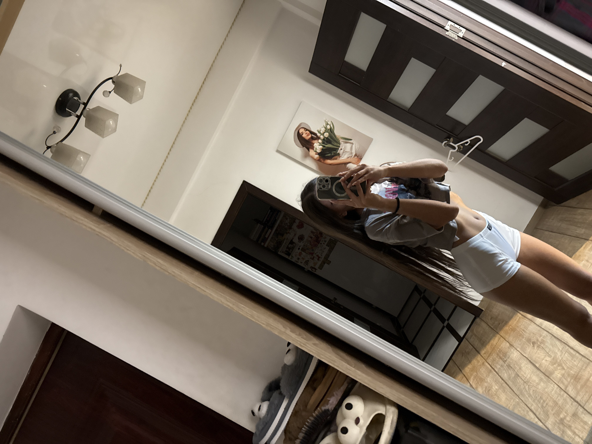 A person is taking a selfie in a mirror. They are wearing a grey t-shirt and white shorts. The mirror reflects a room with a wooden floor, a dark brown door with frosted glass panels, and a framed picture of a woman holding flowers on the wall. A ceiling light fixture with three lamps is also visible in the reflection. To the left of the frame, partially visible, are shelves with slippers and a hairbrush. The scene appears to be indoors, likely a bedroom or dressing area, in a residential setting.