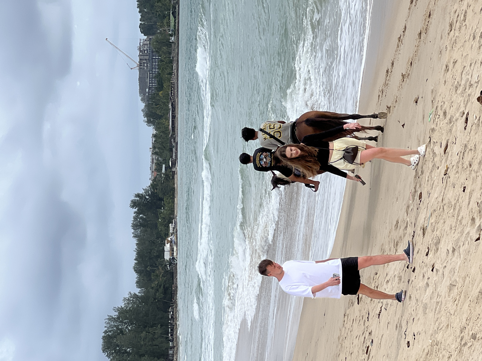 Two young adults walk along a sandy beach with gentle waves lapping at the shore. The man on the left is wearing a white t-shirt and black shorts. The woman on the right is wearing a black long-sleeved shirt and a light-colored skirt. Behind them, three individuals are riding a brown horse in the shallow water. The background features lush green trees and buildings, with a construction crane visible in the distance. The sky is overcast.