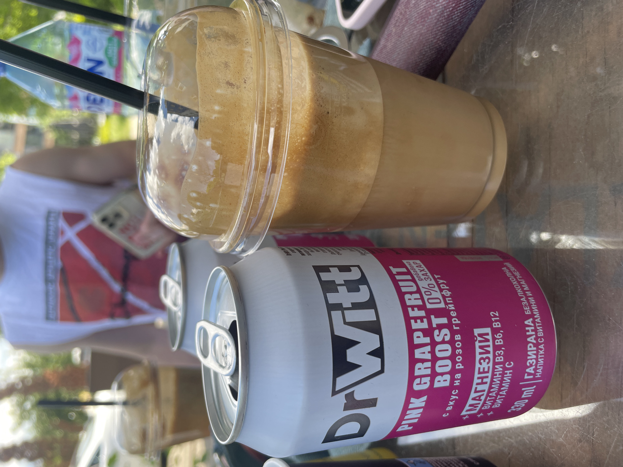 The image shows an outdoor scene, possibly in a cafe or similar establishment. A clear plastic cup filled with a layered beverage, likely an iced coffee or similar drink with a straw, is the main focus. A partially visible can of "Dr. Witt PINK GRAPEFRUIT BOOST 0%" soda is positioned below the coffee. The can is mostly white with pink and black lettering, and it appears to be opened. Another can of the same soda is visible in the background, partially obscured.
A person wearing a white shirt with a red design is partially visible in the background to the left, holding a smartphone. The surface on which the items are placed appears to be a weathered or aged table. Given the Cyrillic text on the soda can, the location is likely in a country that uses the Cyrillic alphabet, which includes Bulgaria. Given the can name and the prevalence of similar products in Sofia, this scene is likely in Sofia, Bulgaria.