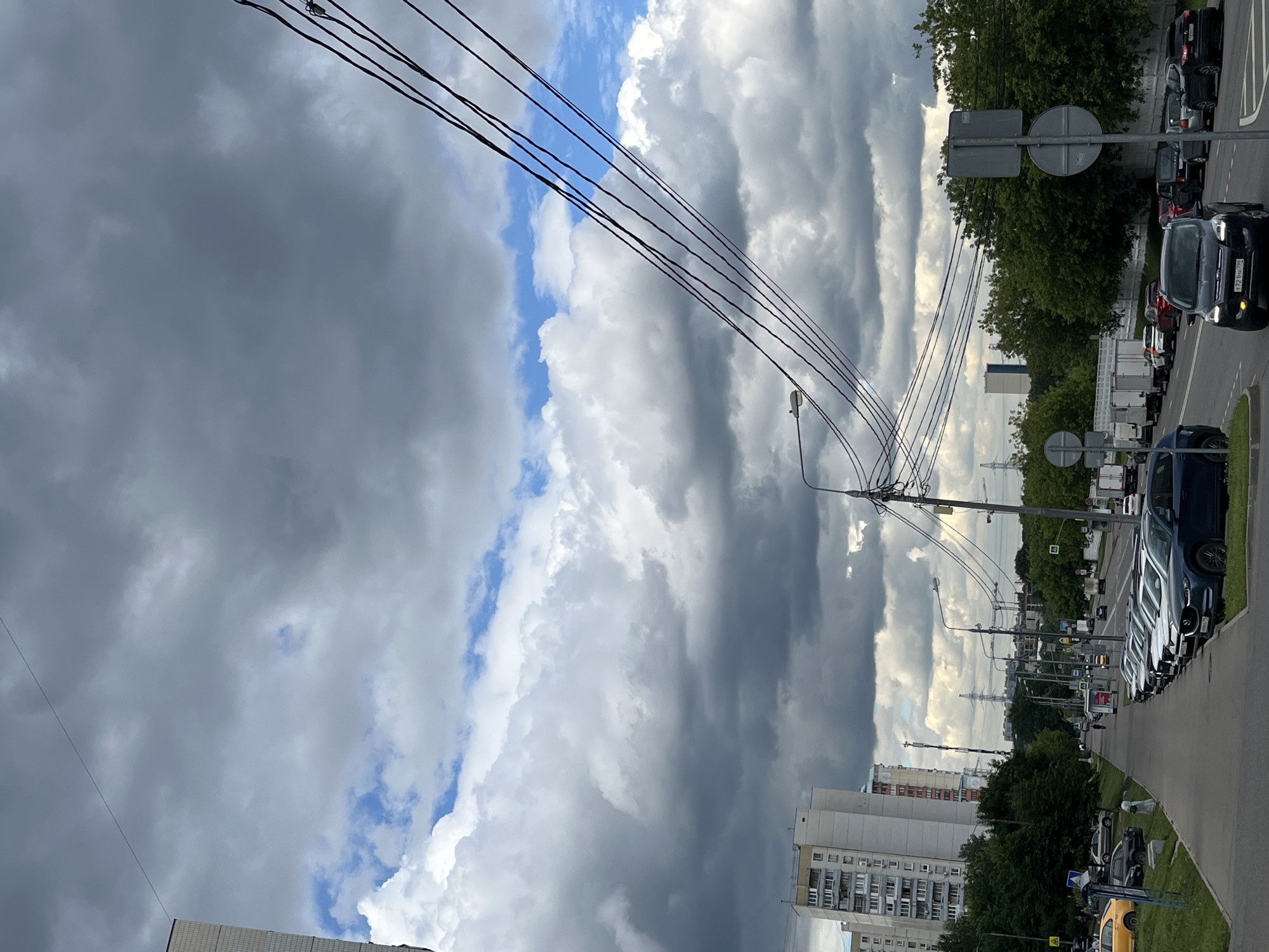 The image shows a street scene in Moscow, Russia. A line of cars is parked along the right side of a road, facing away from the viewer. Overhead, a tangled network of electrical wires stretches across the frame. In the background, tall buildings and trees are visible, with a cloudy sky overhead. The clouds are a mix of dark gray and white, suggesting an overcast or stormy day. The lighting indicates it is daytime, but the exact time is not specified. There is no visible text in the image. The scene appears to be a typical urban environment.