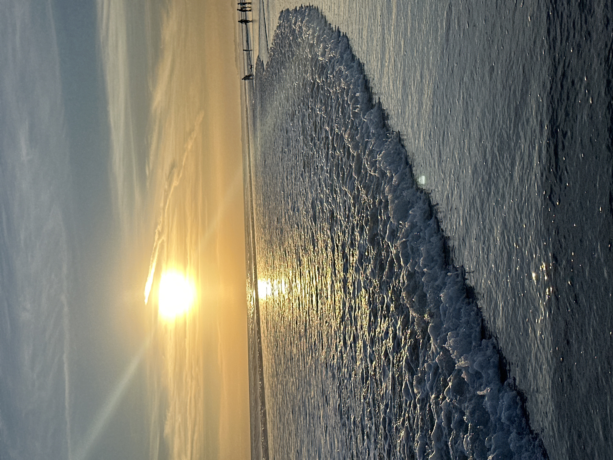 Captured at an angle, the image portrays a beach scene, likely in North Kuta, Indonesia, given the presence of figures on the shore. The sun, positioned low in the sky, casts a bright, golden glow, creating a reflection on the ocean's surface. The water is turbulent, with visible waves and foam. Along the shoreline, there are a few indistinct figures, potentially people engaging in activities. The sky is filled with wispy clouds.