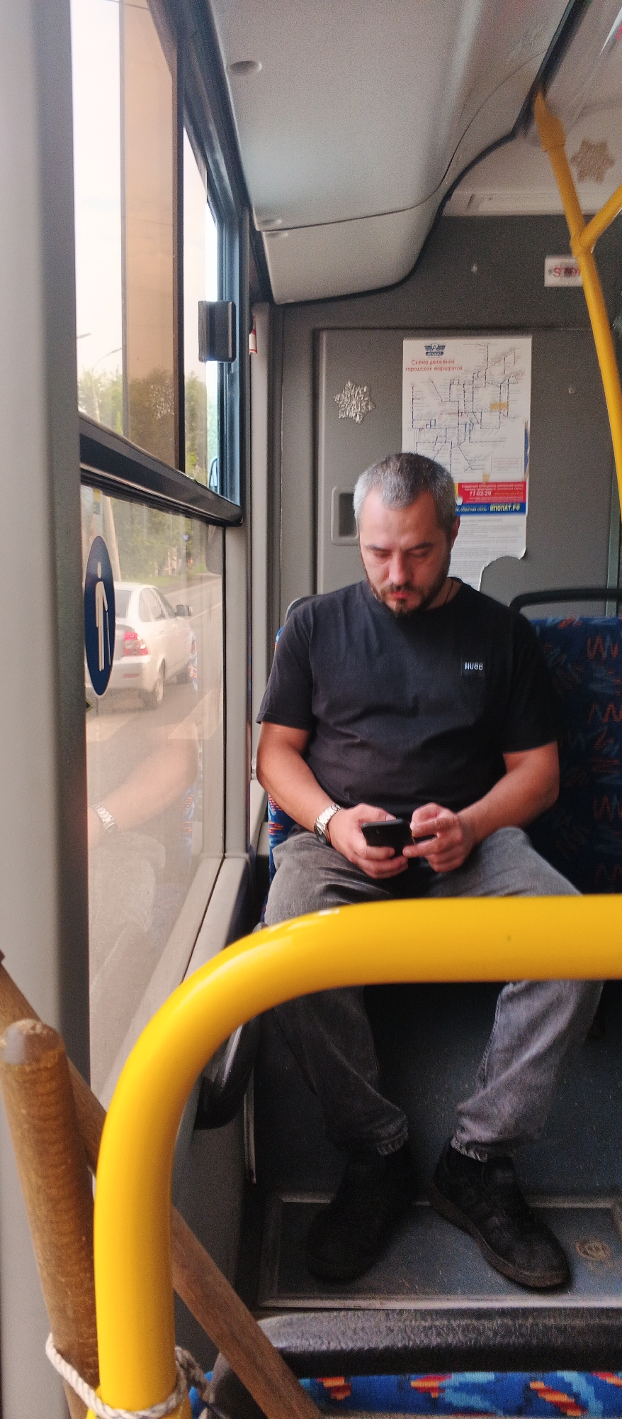 A man with graying hair and a beard is seated inside a bus, looking down at his smartphone. He is wearing a black t-shirt with a "HUGO" logo and gray jeans. His right wrist sports a silver watch. The bus interior features blue and orange patterned seats and a yellow safety bar in the foreground. Through the window, a white car is visible on a road bordered by greenery, suggesting an urban or suburban setting. A map of city routes is affixed to the wall behind him. The image appears to be taken in Izhevsk, Russia, given the context provided.