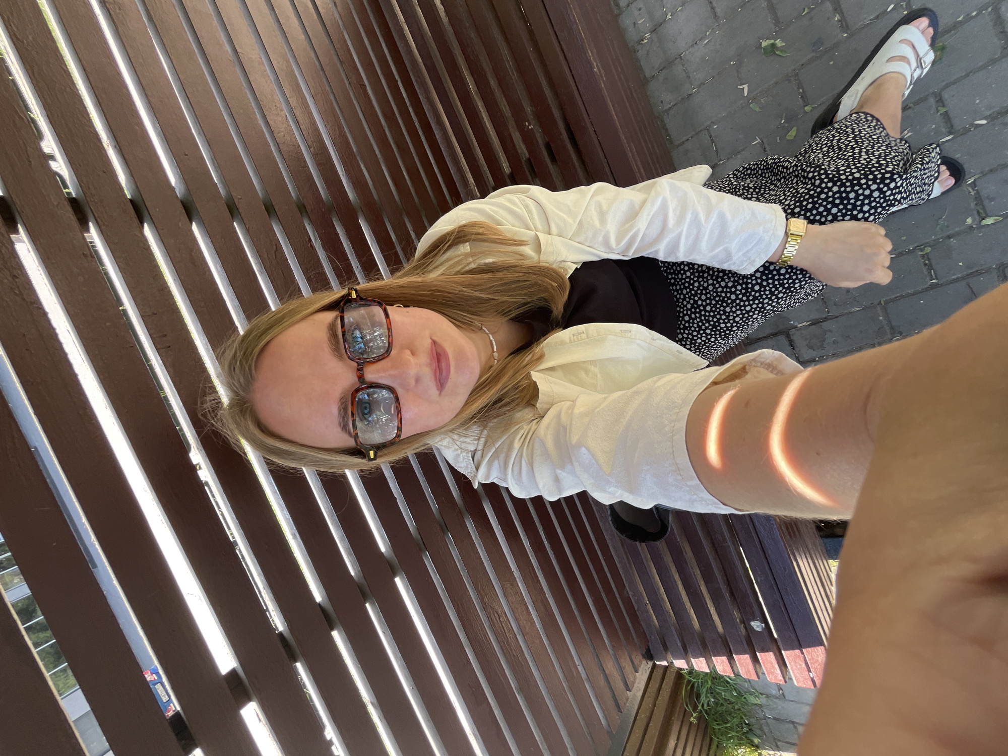A high-angle, medium shot captures a young woman with blonde hair and glasses sitting on a wooden slatted bench outdoors, holding her phone to take a selfie. She wears a light-colored jacket over a dark top and a black and white polka dot skirt. A gold watch is visible on her left wrist, and she has on white sandals. The bench is positioned in an outdoor urban setting, with a paved ground area visible below. Sunlight casts shadows across the scene, dappling the bench and the woman's arm. The background is partially obscured by the bench's slats, but a glimpse of greenery and what appear to be road signs suggests a location in the city of Chaikovsky, Russia.