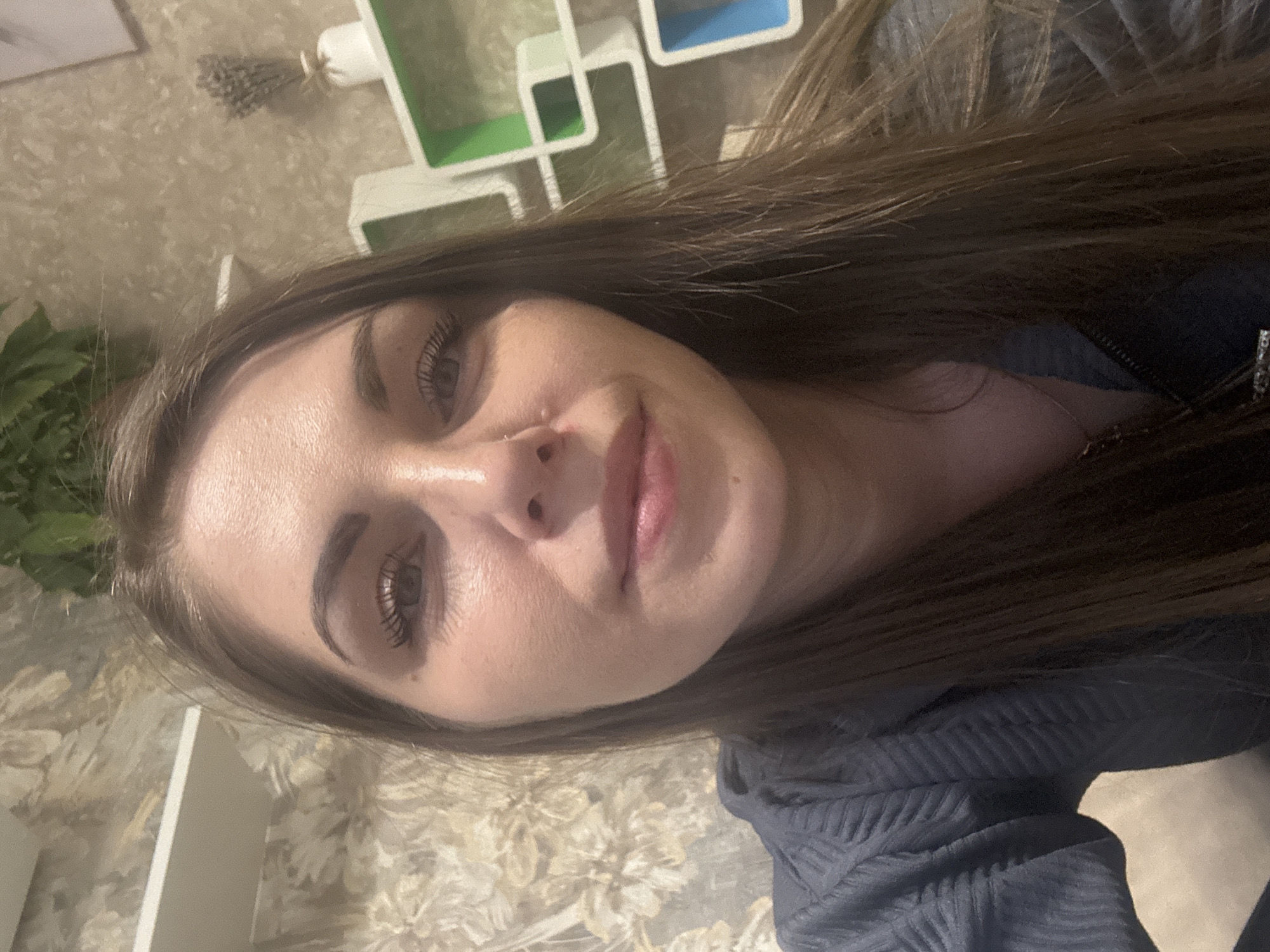 A close-up selfie of a woman with long brown hair and light skin is shown. She is wearing a dark blue or gray textured top. Her gaze is directed slightly off-center from the camera. Behind her, there's a wall decorated with a floral pattern and shelves containing green, white, and blue accents. A bunch of lavender is also visible in the background. There is no information in the image to indicate this location is Раздолье, Russia.
