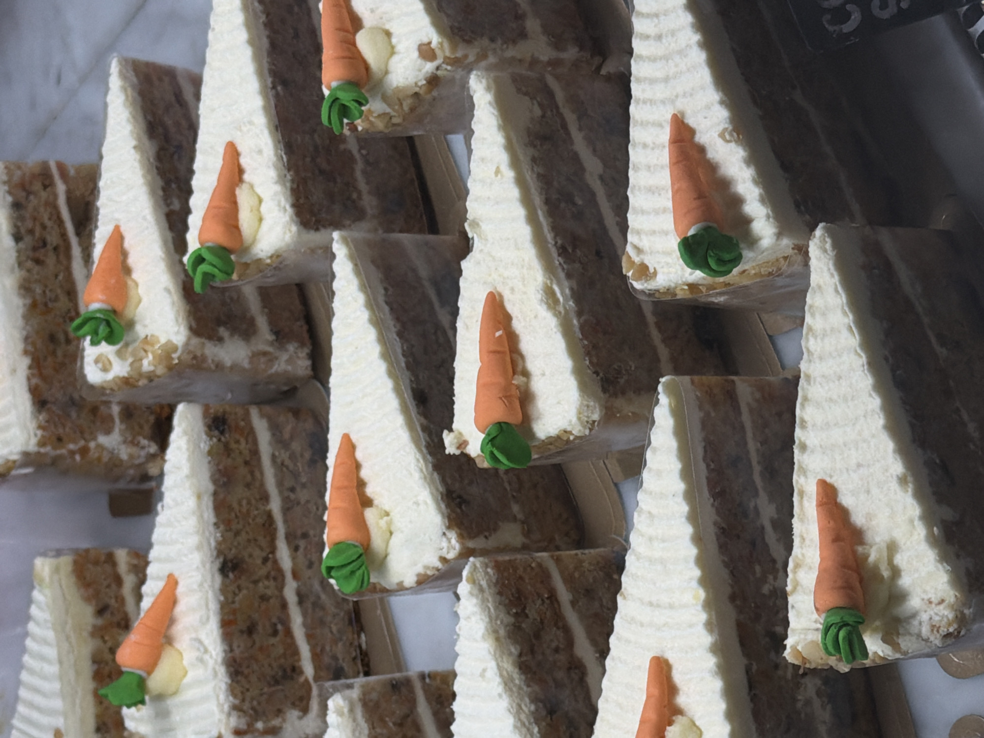 The image shows a display of triangle-shaped slices of carrot cake topped with white frosting and a small, decorative marzipan carrot. The cakes appear to be layered with multiple layers of cake and frosting. The slices are arranged in a stacked, layered fashion. The context of "North Kuta, Indonesia" is not visible within the provided image.
