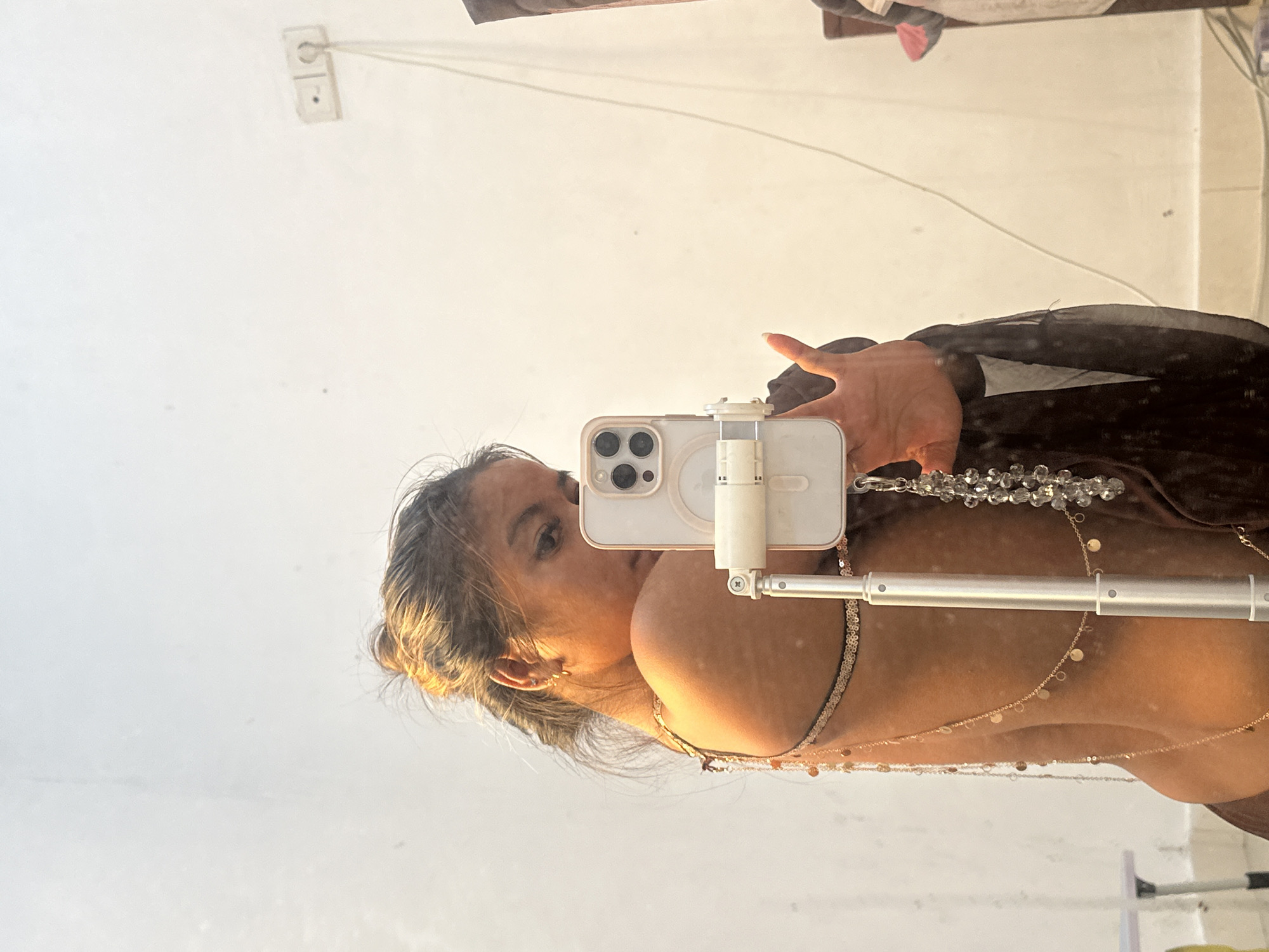A woman is taking a mirror selfie with a white iPhone 14 Pro using a selfie stick. She is wearing a dark brown garment and several gold necklaces. The background is a white wall with an electrical outlet. There is a reflection of other objects on top of the mirror. It's impossible to verify the location is Celuk, Indonesia, as there are no features to confirm it.