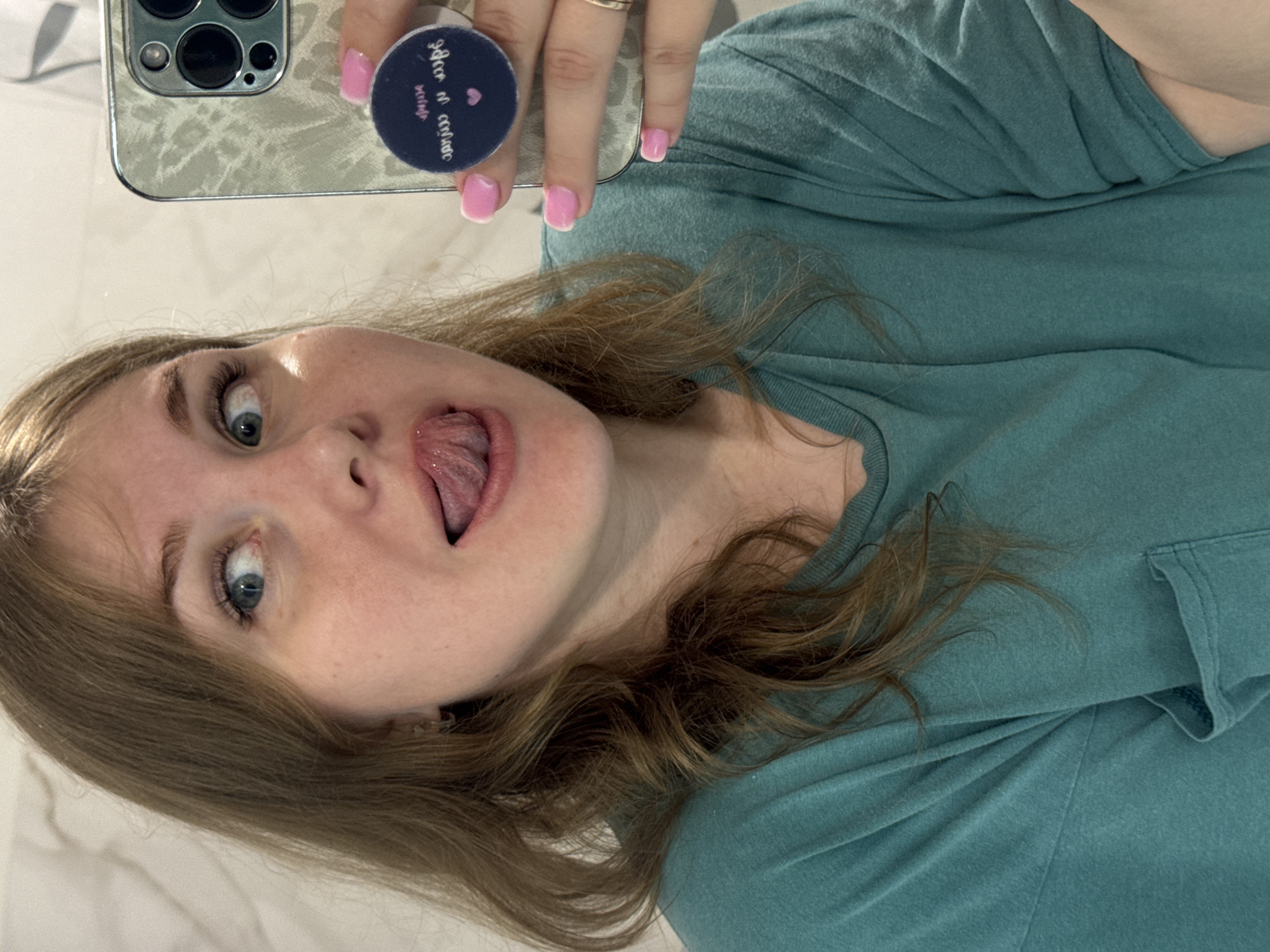 A woman with light brown hair and blue eyes is holding a smartphone in a case up to take a selfie. She is wearing a teal t-shirt and sticking her tongue out. Her nails are painted light pink with white tips. The background appears to be a white, possibly tiled surface. The location, Ферма, Russia, cannot be confirmed based on the image.