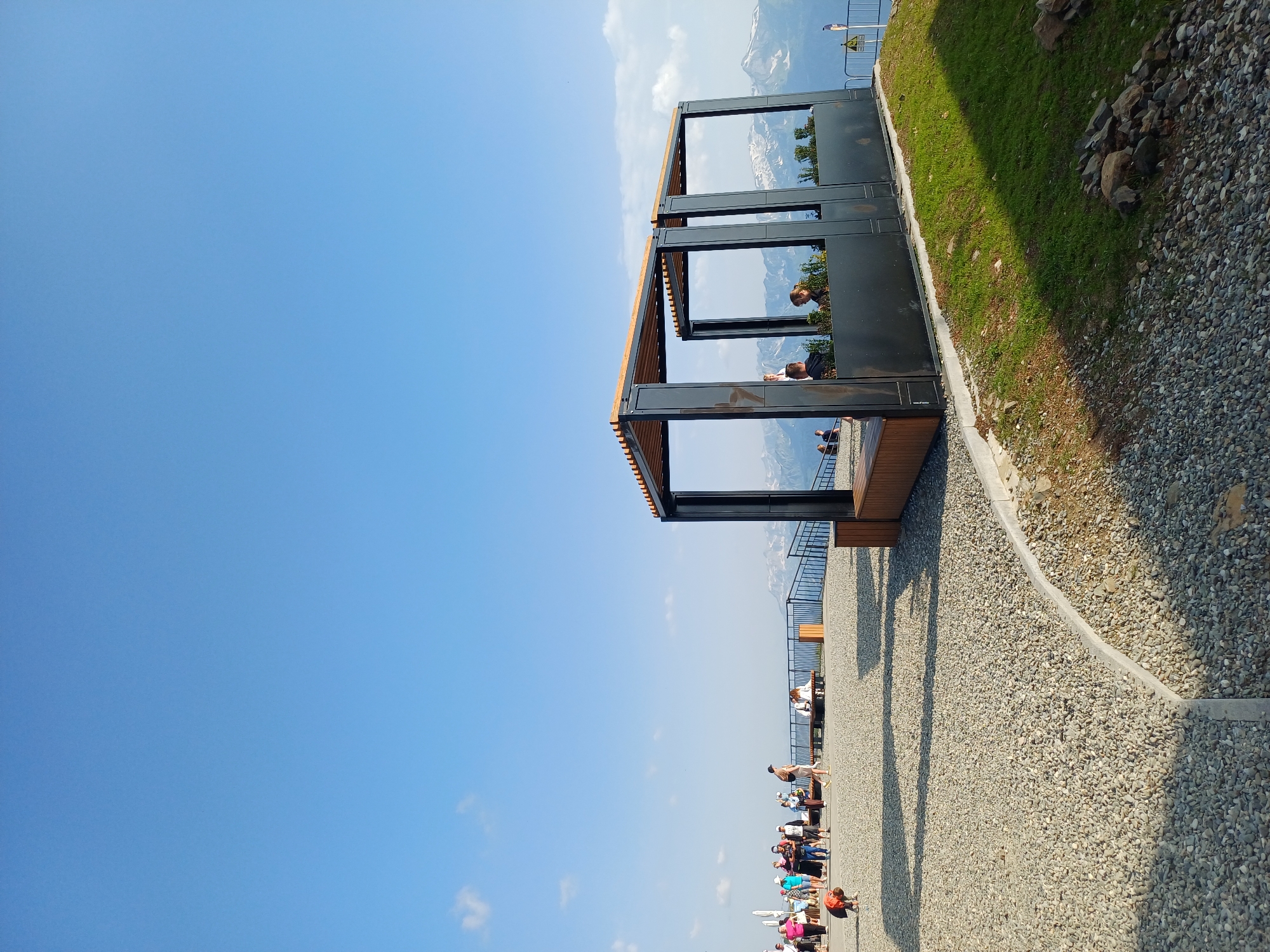 This is a wide-angle, eye-level outdoor photograph of a mountain observation deck on a clear, sunny day. The foreground is a wide, paved area covered in light-colored gravel, with a curved concrete border in the lower left corner. To the right, a grassy slope leads down from the paved area. In the middle ground, there's a modern, open-sided structure with a wooden slatted roof and dark metal framing, appearing to be a seating area or shelter. People are visible on the observation deck, some standing and walking, and others seated within or near the structure. In the background, a metal railing separates the deck from a view of snow-capped mountains under a bright blue sky with a few wispy clouds. The scene appears to be a tourist or recreational area, likely in a mountain resort. The specific location is identified as the observation deck in Estosadok, Russia, known for its mountain views.