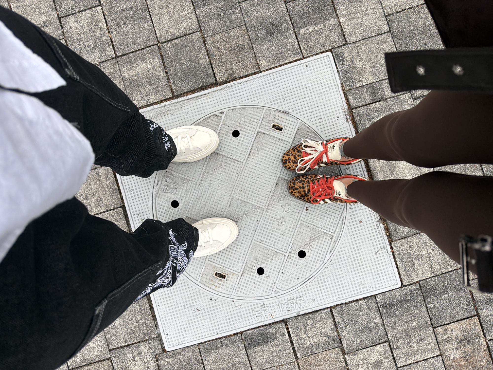 A high-angle shot captures two individuals standing on a manhole cover set in a paved walkway. The person on the left is wearing loose-fitting black jeans with white dragon embroidery along the outer leg, paired with white sneakers. The person on the right is wearing brown leggings and leopard-print Adidas sneakers with red laces. The manhole cover is gray with a patterned surface. The surrounding pavement is composed of gray, square tiles. This image was taken in Gimpo-si, South Korea.