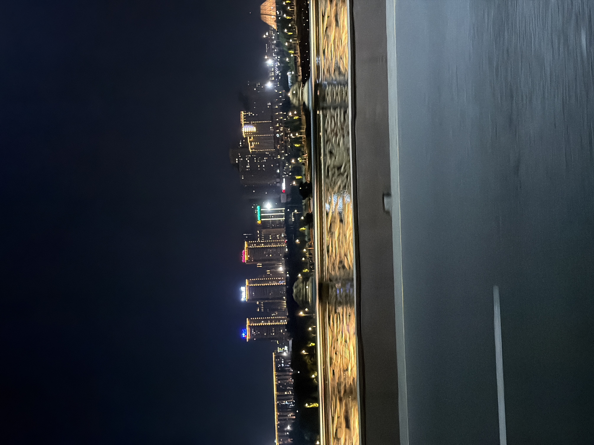 The image captures a nighttime cityscape of Astana, Kazakhstan, viewed from a moving vehicle, likely a bridge. The foreground is dominated by the dark asphalt of a road with a single white lane marker, indicating motion. Beyond the road, a railing is visible, separating it from a body of water. The water reflects the city lights, creating streaks of yellow and orange that shimmer on the surface.
In the mid-ground, a waterfront promenade or park area is discernible, lined with streetlights casting warm pools of illumination. Trees are sparsely dotted throughout this area.
The background showcases the Astana skyline. Several modern high-rise buildings are illuminated with strings of lights, some featuring colored accents. Notable structures include a building with a prominent blue light on its upper section and another with a large yellow illuminated "4." Further to the right, a distinctive pyramid-shaped building with a faceted roof is visible, a recognizable landmark of Astana. The sky above is dark and clear.