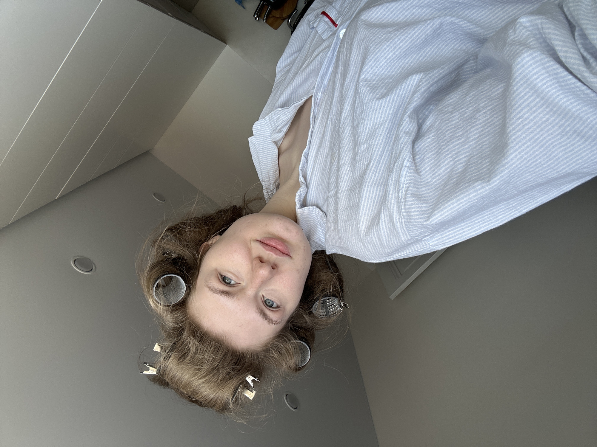 A young Caucasian woman is depicted from a low-angle perspective. She has fair skin, blue eyes, and light brown, curly hair styled with silver hair curlers and clips. She's wearing a light blue and white striped button-down shirt, which is partially unbuttoned. The environment includes a neutral-colored wall, a ceiling with recessed lighting, and a partial view of a white cabinet. The location is likely indoors, but there is no indication of Kazan, Russia.