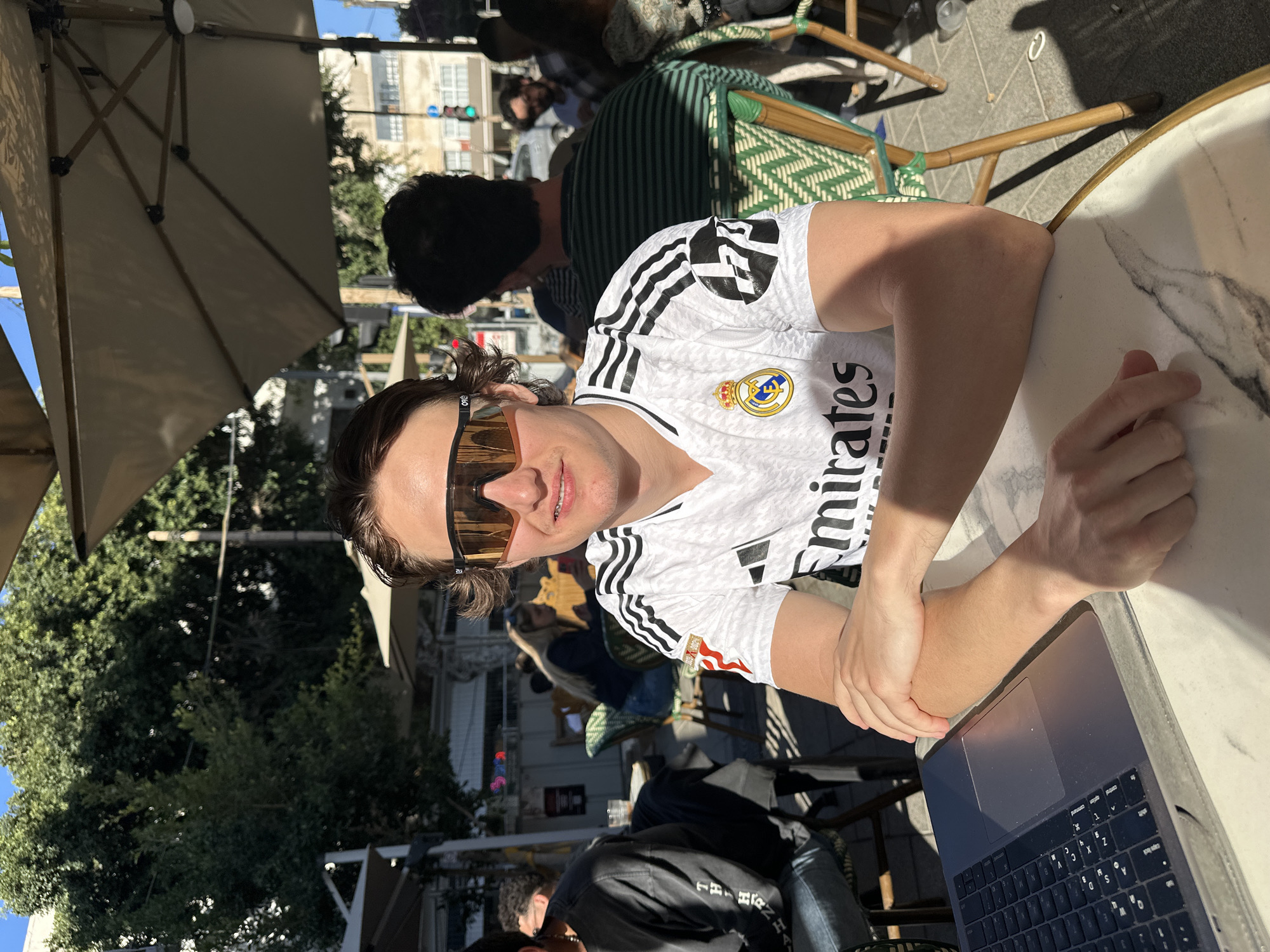 A person is seated at an outdoor cafe table in Tel-Aviv, looking towards the camera with a slight smile. They are wearing a white short-sleeved Real Madrid football jersey featuring black Adidas stripes on the shoulders, the Real Madrid crest, and the sponsor "Emirates" prominently displayed on the front. Large, wraparound sunglasses with orange-tinted, reflective lenses cover their eyes.
The setting is an outdoor cafe, characterized by a large beige umbrella providing shade and green-patterned wicker chairs. The table in front of the person is white with a grey marble-like pattern. A silver laptop, appearing to be a MacBook, is open on the table to the person's right, showing its keyboard and trackpad. The person's left arm rests on the table, while their right hand is positioned near the laptop.
In the background, other patrons are visible seated at various tables, some also under umbrellas. Beyond the cafe seating, city buildings, abundant green trees, and a traffic light displaying a green signal are visible under a clear, bright blue sky. The scene is brightly lit by natural sunlight, indicating a sunny day, with distinct shadows cast across the tables and ground. The overall atmosphere suggests a relaxed urban environment.
