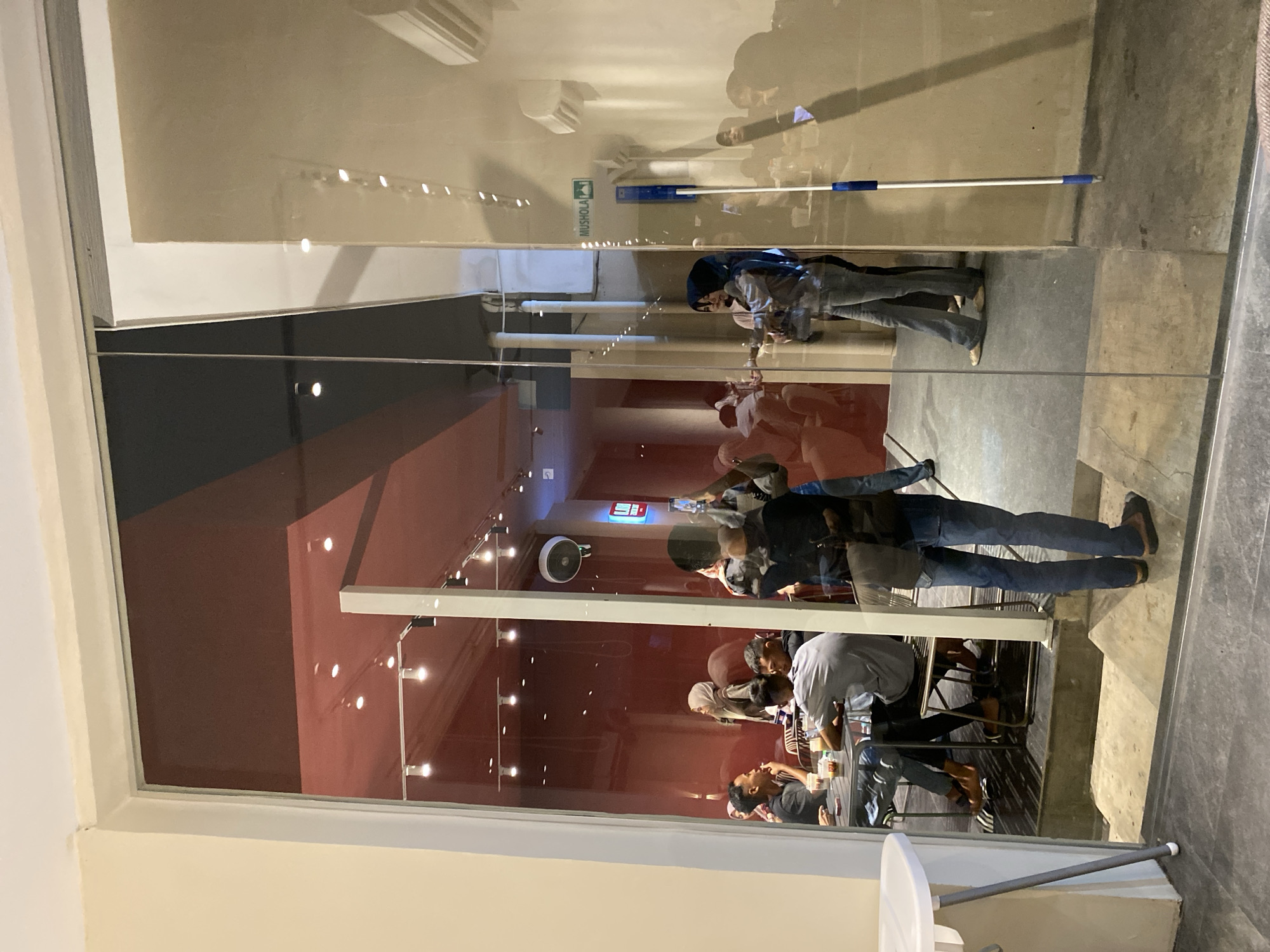The image captures a scene within a cafe in Lhokseumawe, Indonesia, viewed through a glass window. Inside, several individuals are seated at tables, some engaged in conversation and others using mobile phones. The interior features red walls and ceiling lights. The exterior shows the sidewalk and a glimpse of the surrounding environment, including a sign reading "Mushola". Reflected on the glass are overhead lights and additional street elements.