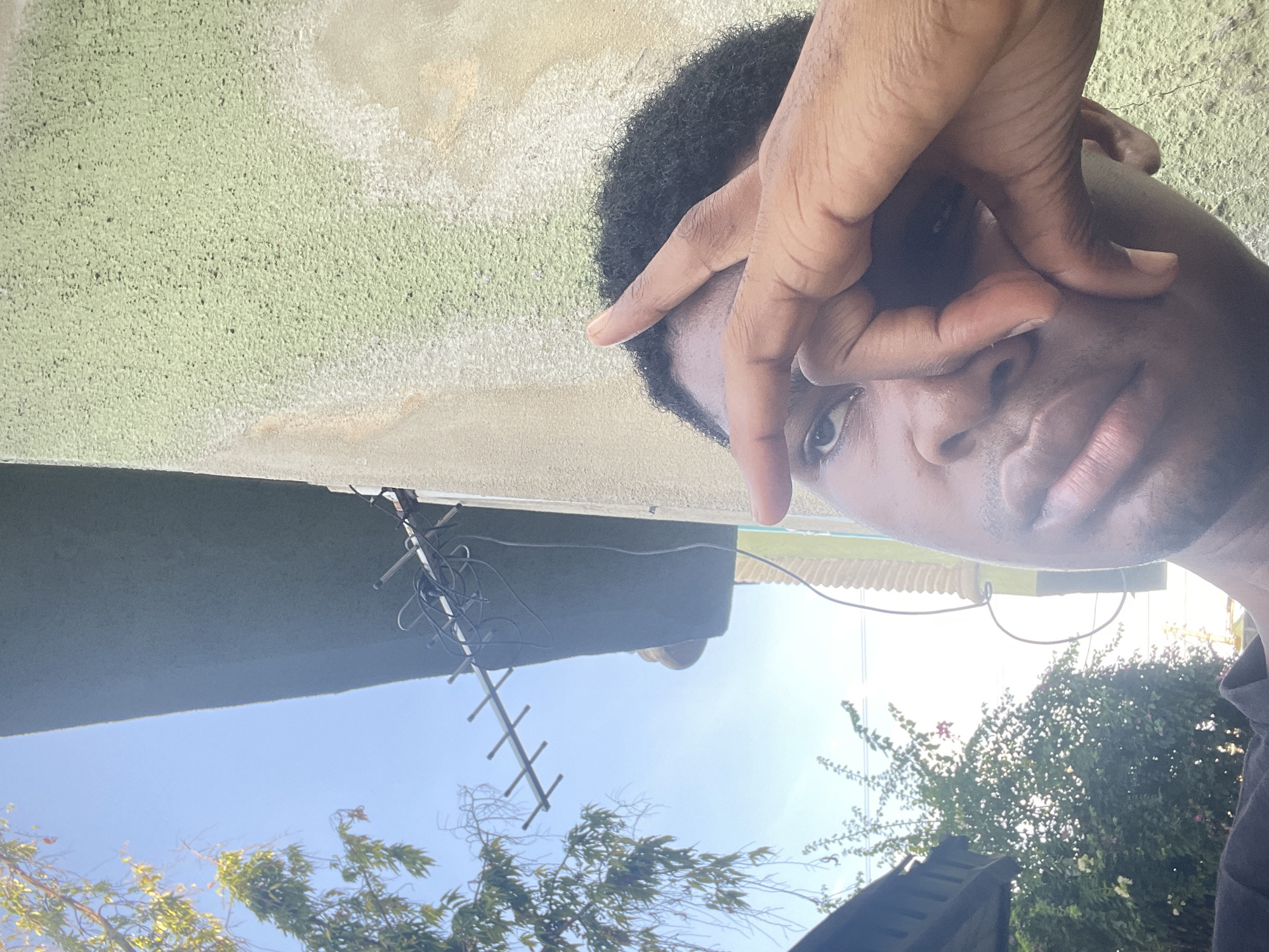 A close-up, tilted selfie features a dark-skinned man with short, curly hair, partially obscuring his face with his hand. His right eye is visible, and his lips are partially closed. Overhead, a light green textured wall is visible. Attached to the wall is a TV antenna with visible wires. Outside, the sky is blue, and the tops of trees with green foliage are visible. This image was taken outdoors and has no visible addresses or distinguishing objects to determine the exact location in Ado, Nigeria.
