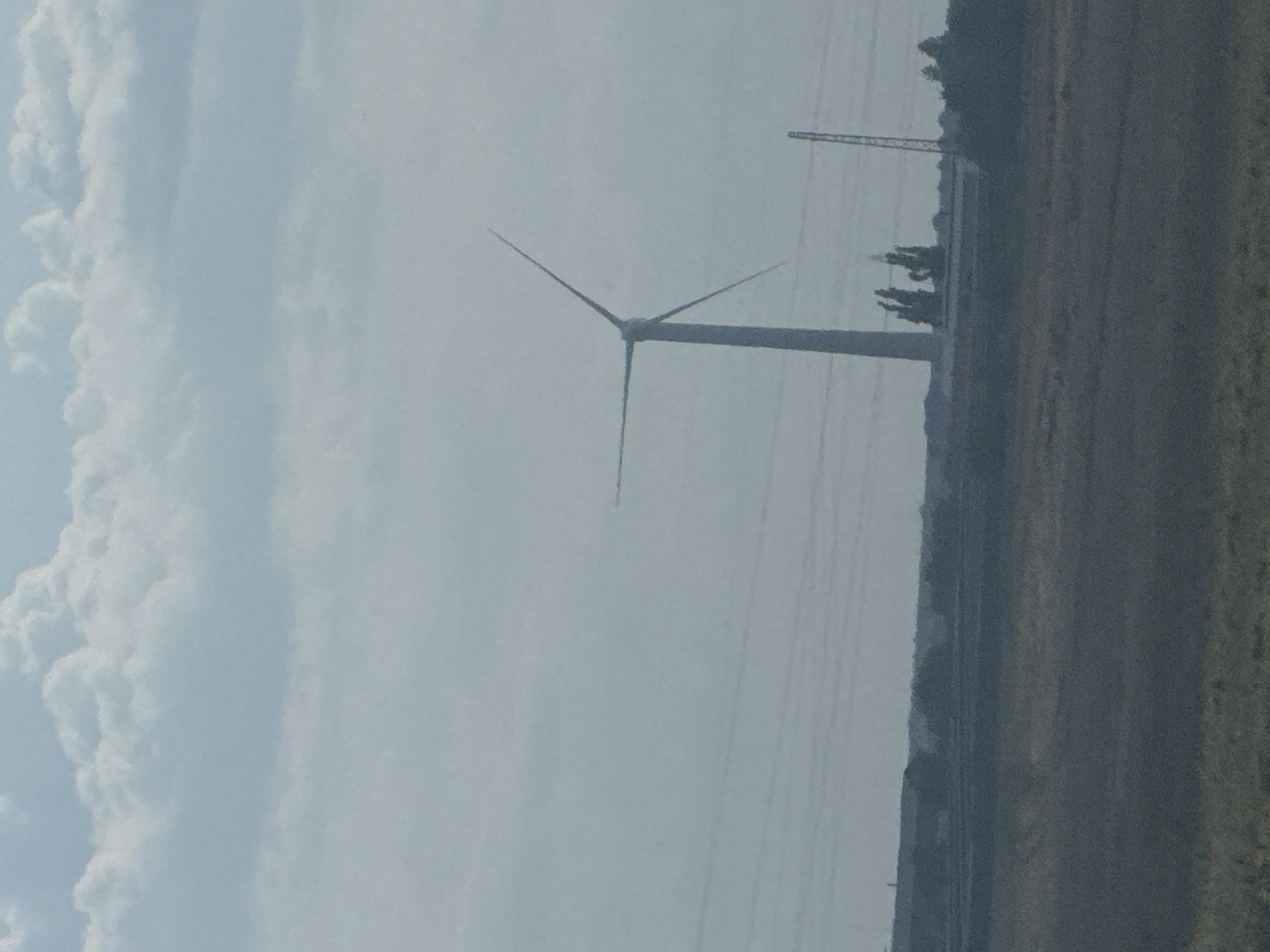 An outdoor, slightly overcast scene features a wind turbine prominently in the center, with its blades angled. In the distance, a line of trees and industrial structures is visible, with a thin, tall tower to the right of the wind turbine. The foreground consists of a grassy, brownish field. The overall impression is one of a rural or semi-industrial landscape under a cloudy sky.