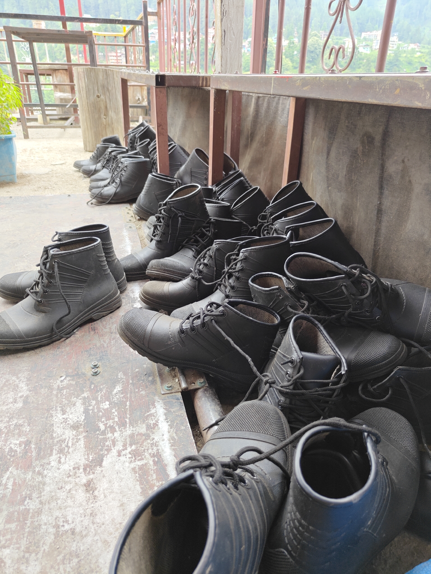 The image displays a multitude of black, lace-up boots, likely made of rubber or a similar waterproof material, arranged in rows and piles on a worn, light-colored platform. Many pairs are stacked against a low wall or barrier on the right side of the frame, while several others are neatly lined up on the left foreground. These boots appear sturdy and practical, possibly designed for trekking or wet conditions.
The setting is an outdoor or semi-outdoor area, possibly a covered veranda or a gear station. A brown metal railing with decorative scrollwork runs horizontally across the mid-ground, providing a partial barrier. Beyond this railing, a lush, green, mountainous landscape is visible, with hints of buildings nestled among the trees, consistent with the location context of Pulga, India. To the left, a large, rustic wooden pillar or log stands upright, and a light blue pot with green foliage adds a touch of plant life.
No specific activity or event is directly captured, but the quantity and type of boots strongly suggest preparation for or conclusion of an outdoor activity, such as hiking or trekking, where such footwear would be essential. The natural, diffused lighting indicates it is daytime, possibly under an overcast sky or during late morning/early afternoon. There is no visible text in the image.