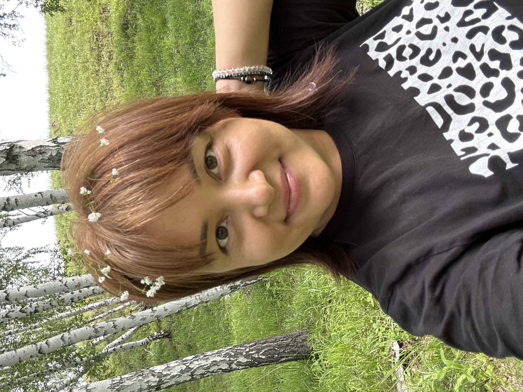 A mid-shot, outdoor, selfie-style photograph shows a woman with shoulder-length, reddish-brown hair adorned with small white flowers, and bangs. She is wearing a black t-shirt with a large, black and white leopard print design on the right side. The woman is smiling and has her right arm raised behind her head. She is wearing two bracelets on her right wrist, one appears to be made of dark beads and the other of clear beads. The background consists of a grassy hill with several birch trees, some with their characteristic white and black bark. The lighting suggests it is daytime, possibly a bright, sunny day. The location is a natural outdoor environment, likely a park or a wooded area, in an unspecified city.