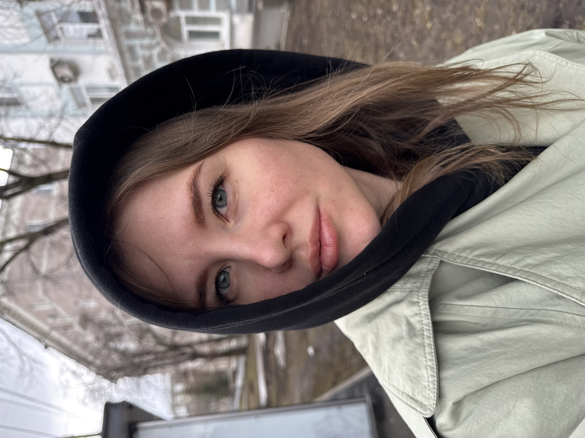A woman is looking at the camera while wearing a black hoodie under a light green jacket. She has light skin and long, light brown hair that is partially obscured by the hoodie. Her eyes are light blue-green. She has a neutral expression.
The background features a blurred urban landscape. In the left background are leafless trees and a light-colored building. In the far background there is possibly another building. The scene appears to be outside during daytime. Based on buildings, window styles, and tree type, the image was likely taken in Russia. Based on the style of the building, Perm, Russia is likely.