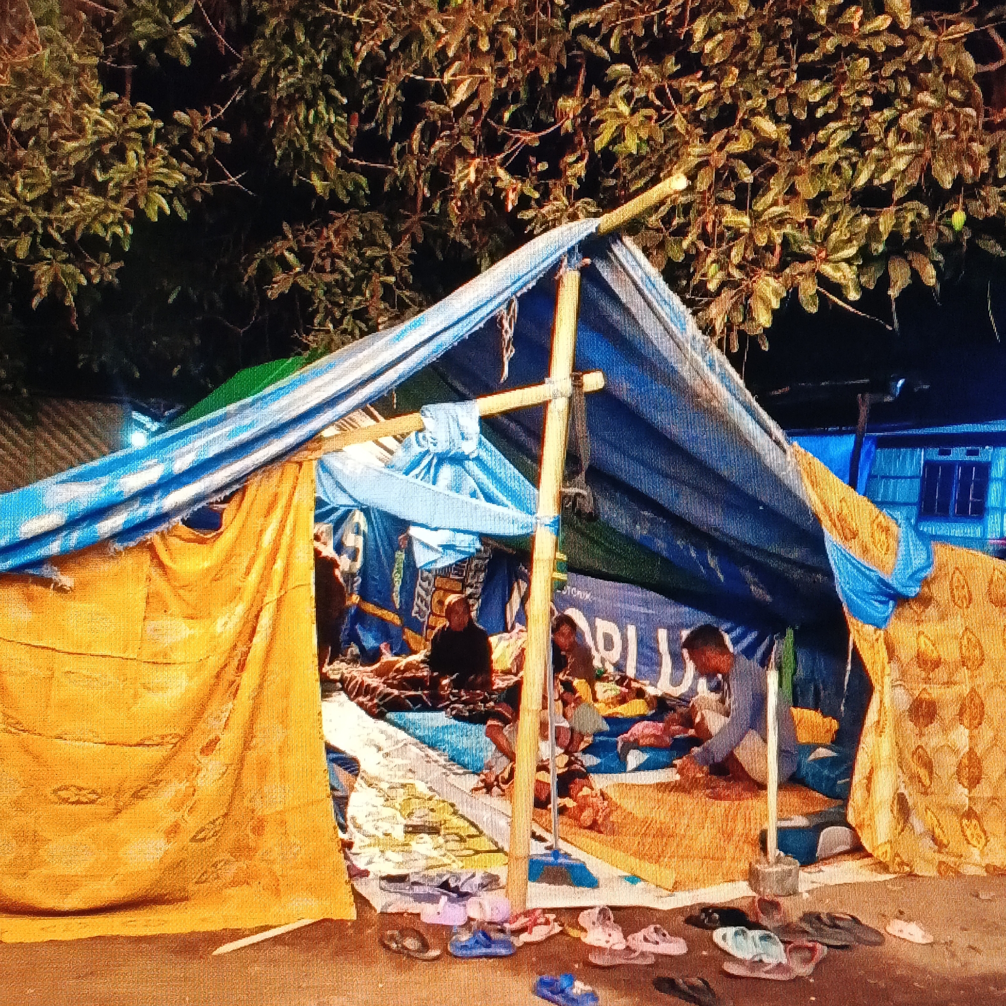 The image depicts a makeshift tent shelter constructed from blue and green tarps and a large yellow patterned cloth, set up outdoors. Several people are visible inside and around the tent. In the foreground, a collection of sandals and flip-flops are scattered on the ground. The tent appears to be a temporary living space, possibly for individuals displaced by an event. The background shows foliage from trees overhead and a building with blue walls and windows. The overall scene suggests a humble and informal settlement. The context of the city of Ende, Indonesia, implies this could be a shelter for those affected by local environmental conditions or other circumstances requiring temporary housing.