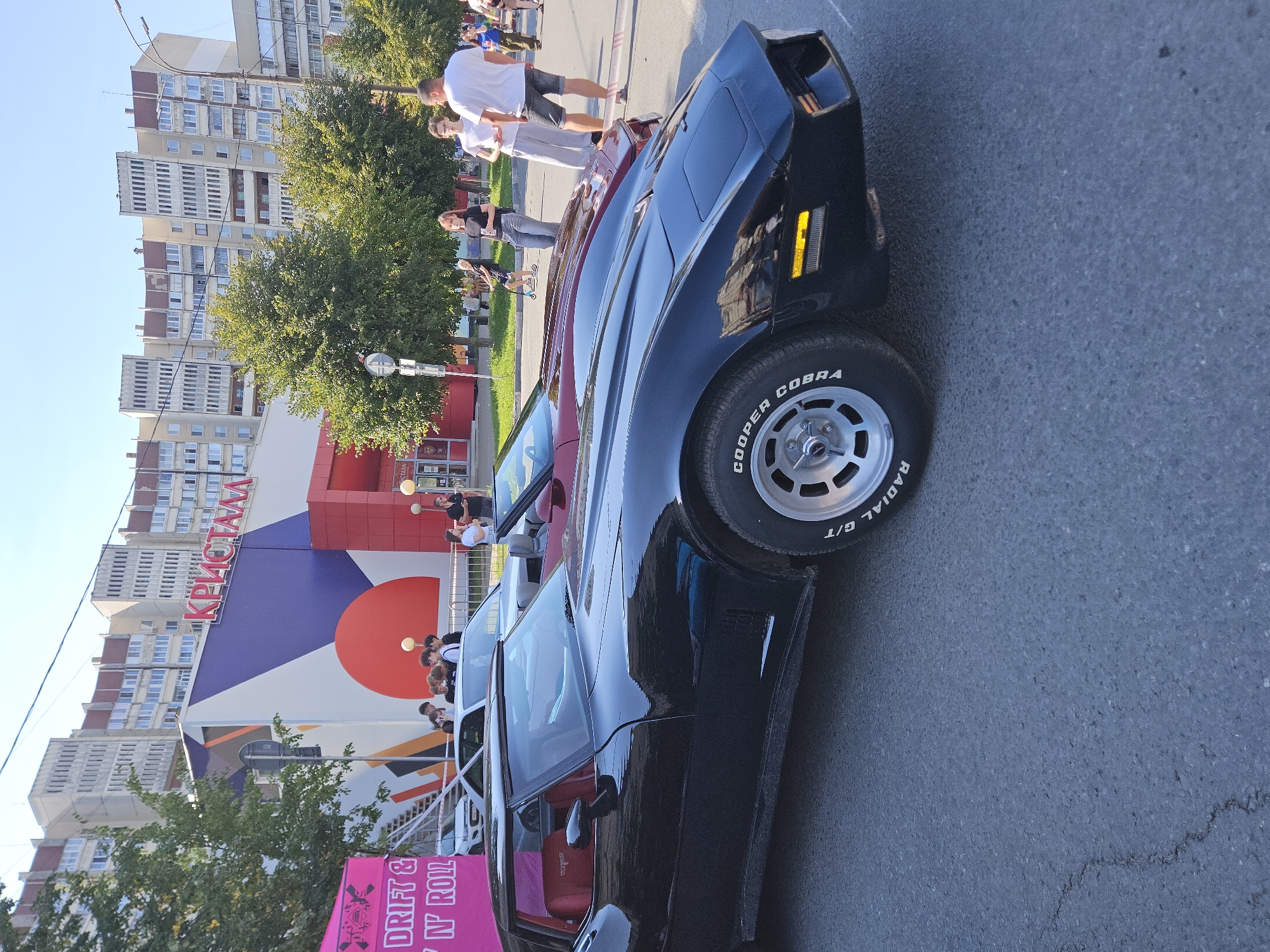 This is a medium shot image taken outdoors on a sunny day, likely in an urban setting in Ulyanovsk, Russia. In the foreground, two classic sports cars are parked side-by-side on an asphalt road. The car on the left is black with a red interior, and the car on the right appears to be a dark red convertible. Both cars have their fronts facing towards the right of the frame. The black car's tire shows the text "COOPER COBRA RADIAL G/T".
Behind the cars, a building with a vibrant, abstract design on its facade is visible. The building features large letters spelling out "КРИСТАЛЛ" (Kristall), suggesting it's a commercial establishment, possibly a shopping center or supermarket. In front of the building, there are several people, some standing and observing the cars, others appearing to be walking or socializing. A bright pink banner with text is also present on the left side of the frame. The background consists of trees and apartment buildings, typical of a Russian city environment. The sky is clear and blue. The scene suggests an outdoor event or gathering centered around vintage cars.