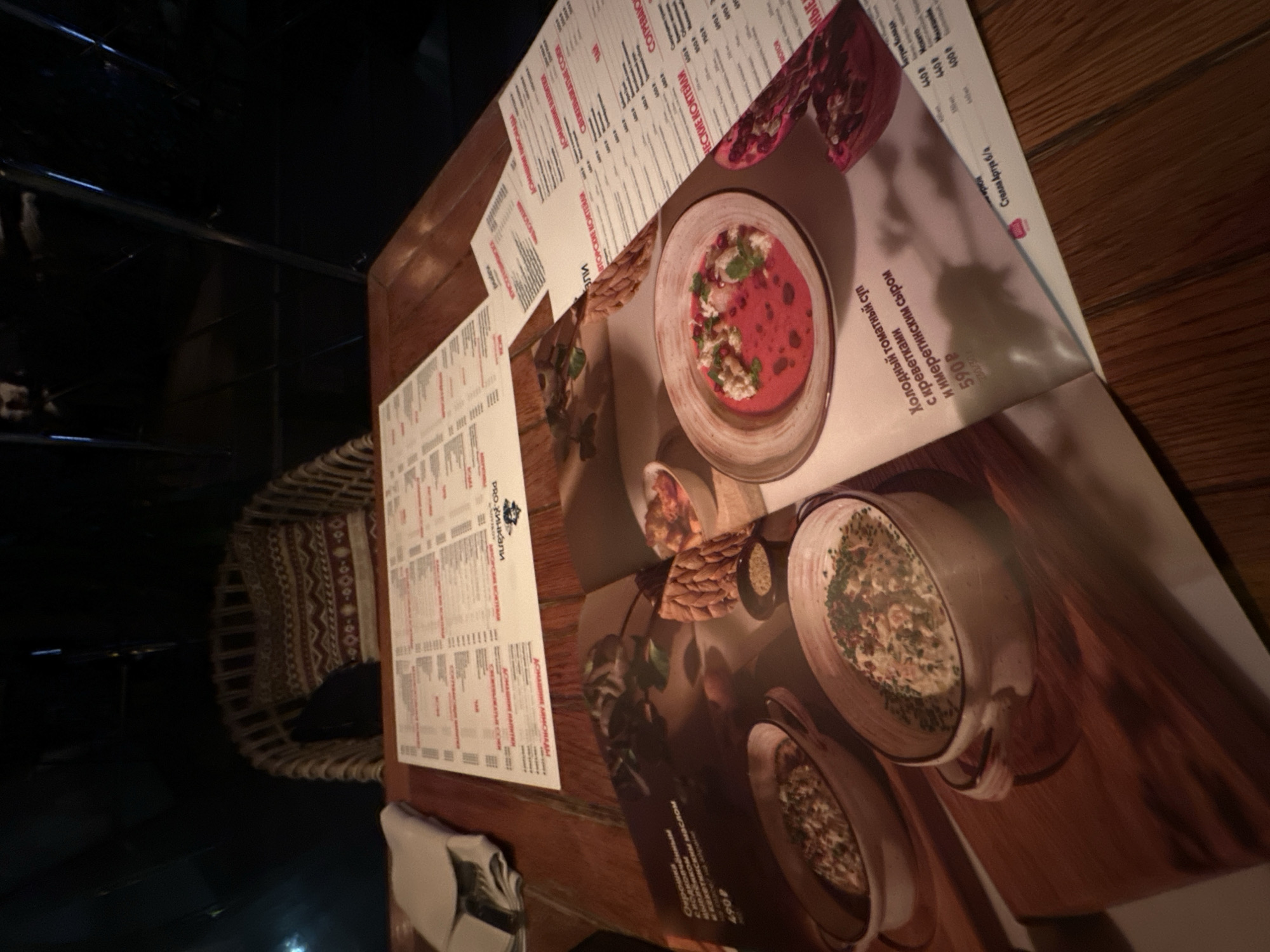 The image depicts a dimly lit restaurant setting, likely in Sochi, Russia, with a focus on a table laid with menus and food. Two open menus are prominently displayed on a wooden table. One menu features appetizing photographs of two dishes served in rustic bowls, one of which appears to be a creamy soup or stew, and the other a vibrant red soup with garnishes. Text, likely descriptions and prices, is visible in Russian below the images. The second, larger menu, lies open in the background, presenting a more extensive list of offerings, also in Russian. A folded white napkin is visible to the left of the menus. In the background, a wicker chair with a patterned cushion is partially visible, suggesting a comfortable seating area. The overall ambiance is warm and inviting, typical of a dining establishment.