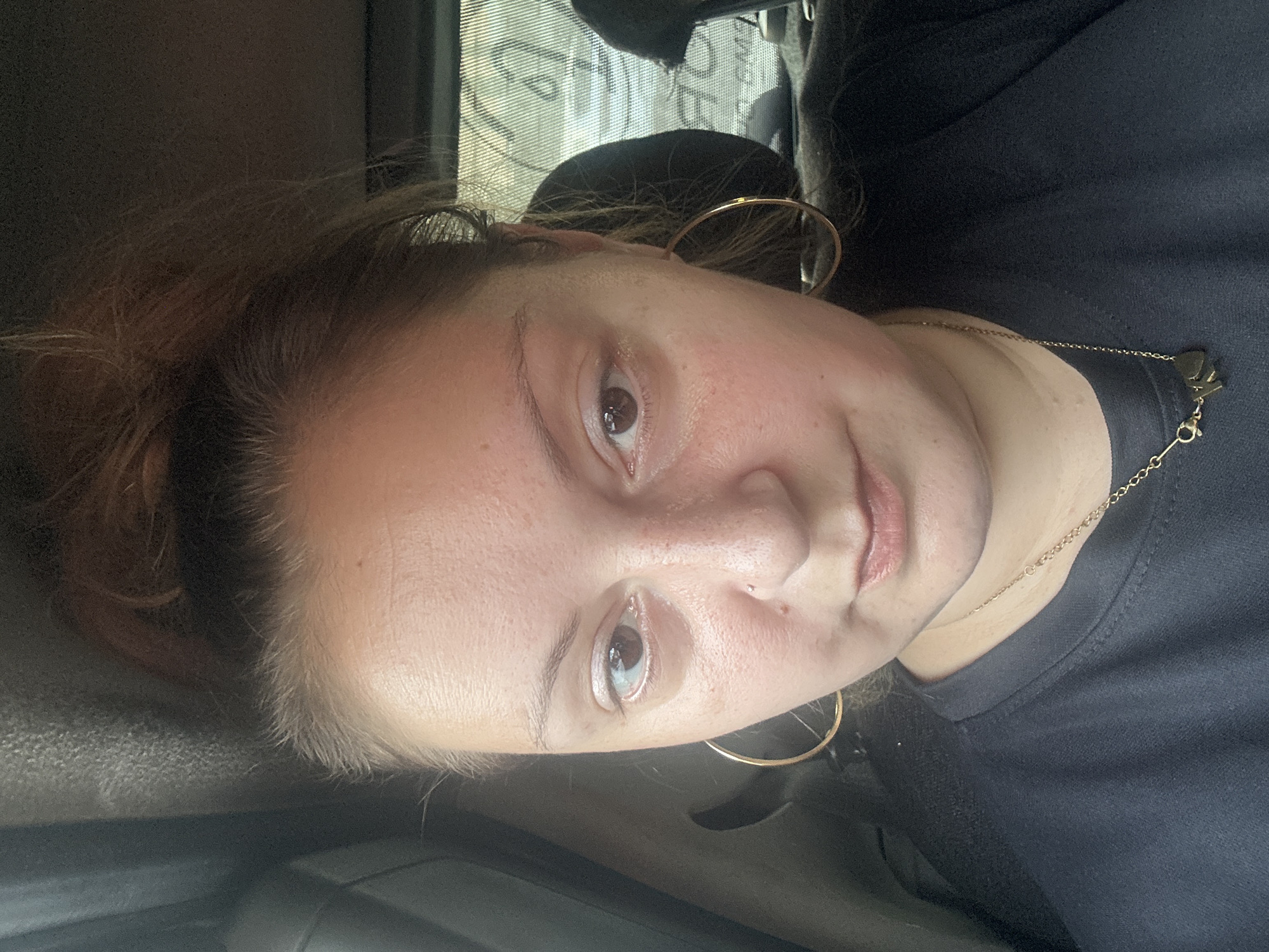 The image is a selfie taken by a young woman with her hair in a bun and wearing a black t-shirt and a gold necklace. She has large hoop earrings and a nose piercing. The background appears to be the interior of a vehicle, with a window visible behind her. The lighting suggests it may be daytime. The scene is devoid of any identifiable landmarks or context that would suggest a location in Tashkent, Uzbekistan.