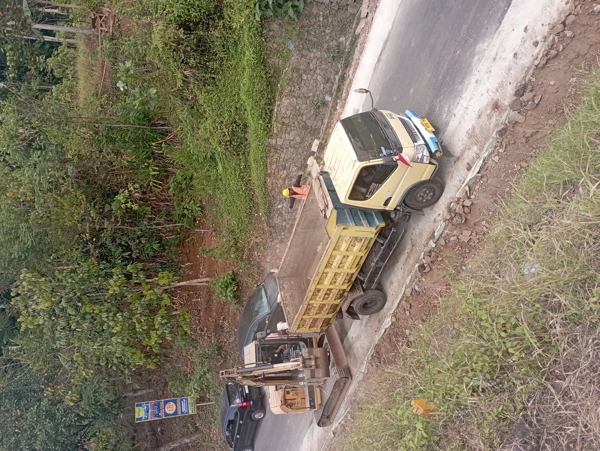 In Manonjaya, Indonesia, an excavator is loading dirt into a yellow dump truck. The excavator is positioned on the left side of the image, with its bucket extended into the truck's bed. The dump truck is a light yellow color and is full of dirt. A person wearing a yellow hard hat and an orange safety vest is standing in the back of the dump truck, appearing to direct the loading process. Another person is visible inside the excavator's cabin. Two black cars are parked on the side of the road in the background. The scene appears to be a construction site on the side of a road, with dense vegetation and trees on a hillside in the background. There is a stone wall on the right side of the road.