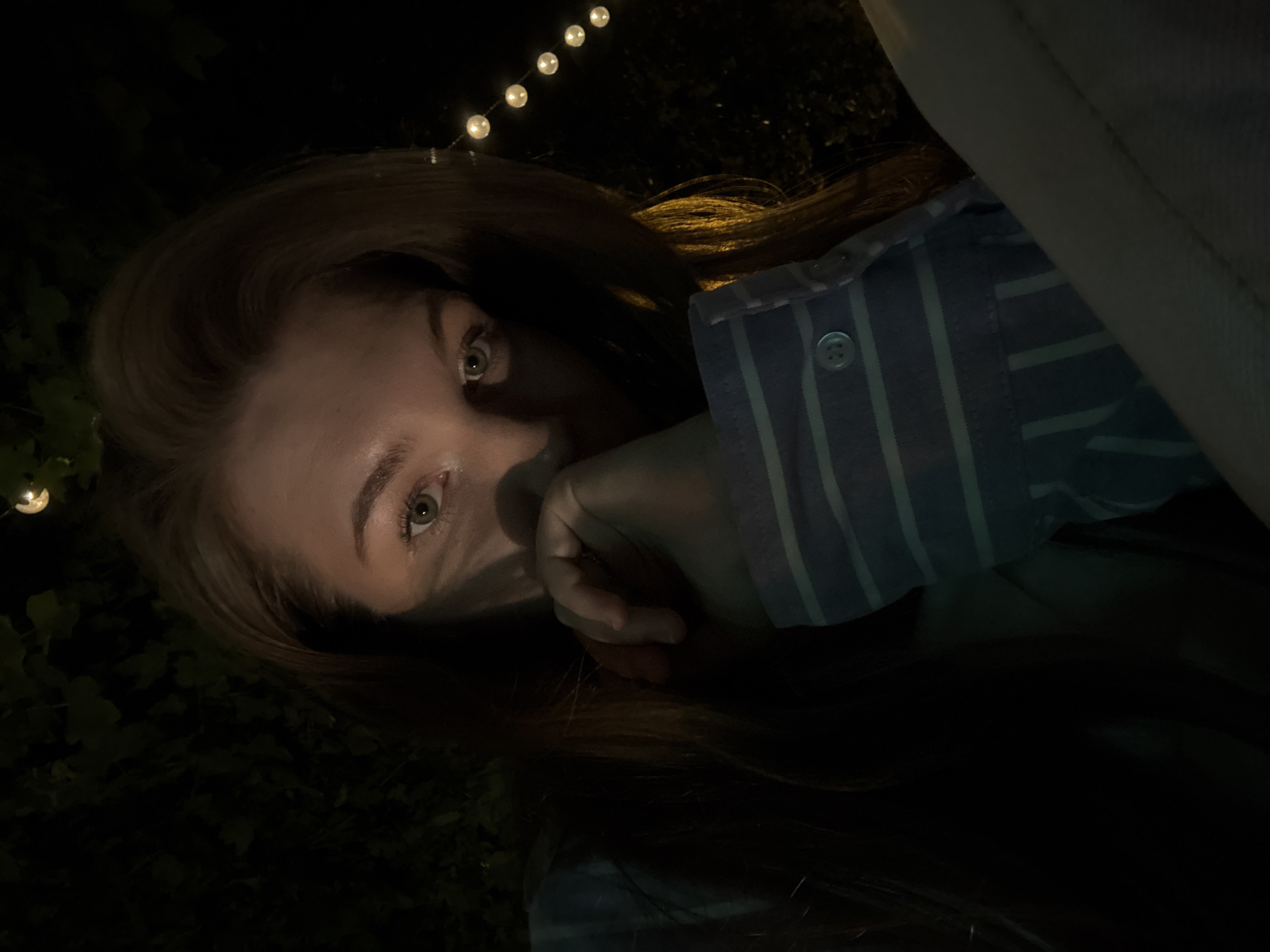 A woman is seen from a low angle in near darkness, with a string of decorative lights visible at the top of the frame. She has fair skin, light brown hair, and light-colored eyes that are directed towards the camera. Her hand is partially covering her mouth. She is wearing a light-colored, vertically striped shirt with the button visible on one of the sleeves. Dark vegetation and possibly ivy can be seen in the background. Location cannot be confirmed as the picture does not have enough identifying information to pinpoint the location within Belgrade, Serbia.