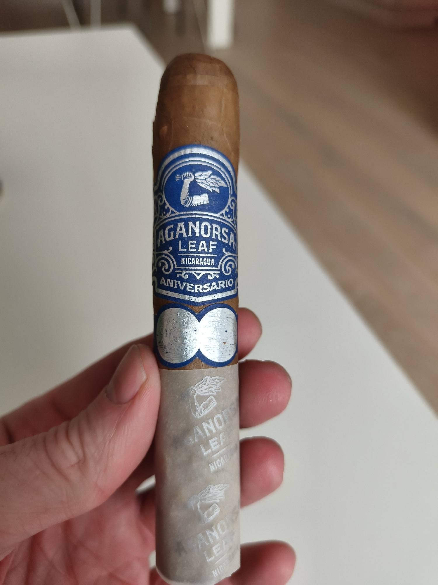 A hand holds a cigar with a blue and silver band labeled "Aganorsa Leaf Nicaragua Aniversario". The cigar is brown and cylindrical. The background shows a light-colored surface. The image does not contain any information that would place it in Sofia, Bulgaria.