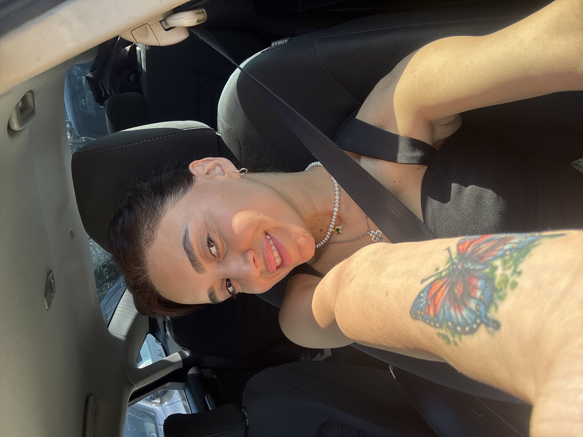 A young woman with brown hair is sitting in the passenger seat of a car. She is smiling and looking at the camera. She has a tattoo of a butterfly on her left arm. She is wearing a black dress and a pearl necklace. The car appears to be parked in an outdoor area.