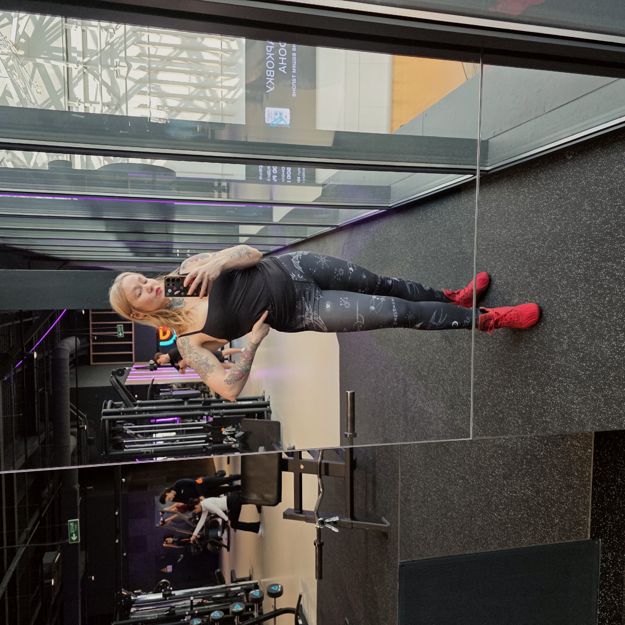 Here is a detailed description of the image:
**Scene:** The image appears to be taken at a gym, based on the presence of exercise equipment visible in the reflection of the mirror and other equipment near the floor. A fair-skinned blonde woman is posing for a mirror selfie, holding a phone. The mirror is likely situated in a gym area.
**People:** The main subject is a woman with blonde hair and tattoos on her arms and neck. She's wearing a black tank top, patterned leggings, and red sneakers. Her arms have rose tattoo patterns. Other people are faintly visible in the gym in the reflection of the mirror.
**Objects:** Prominent objects include the woman's smartphone, black exercise equipment, a mirror, the woman's red sneakers and patterned leggings.
**Environment:** The background includes visible elements of the gym such as exercise equipment. Natural light is coming from a large window.
**Additional details:**
- A sign with Russian text is visible in the reflection of the window.
- The walls and flooring have a dark, neutral color scheme.
- The location might be in the city of Saint Petersburg, Russia, based on the Russian text in the reflection.
