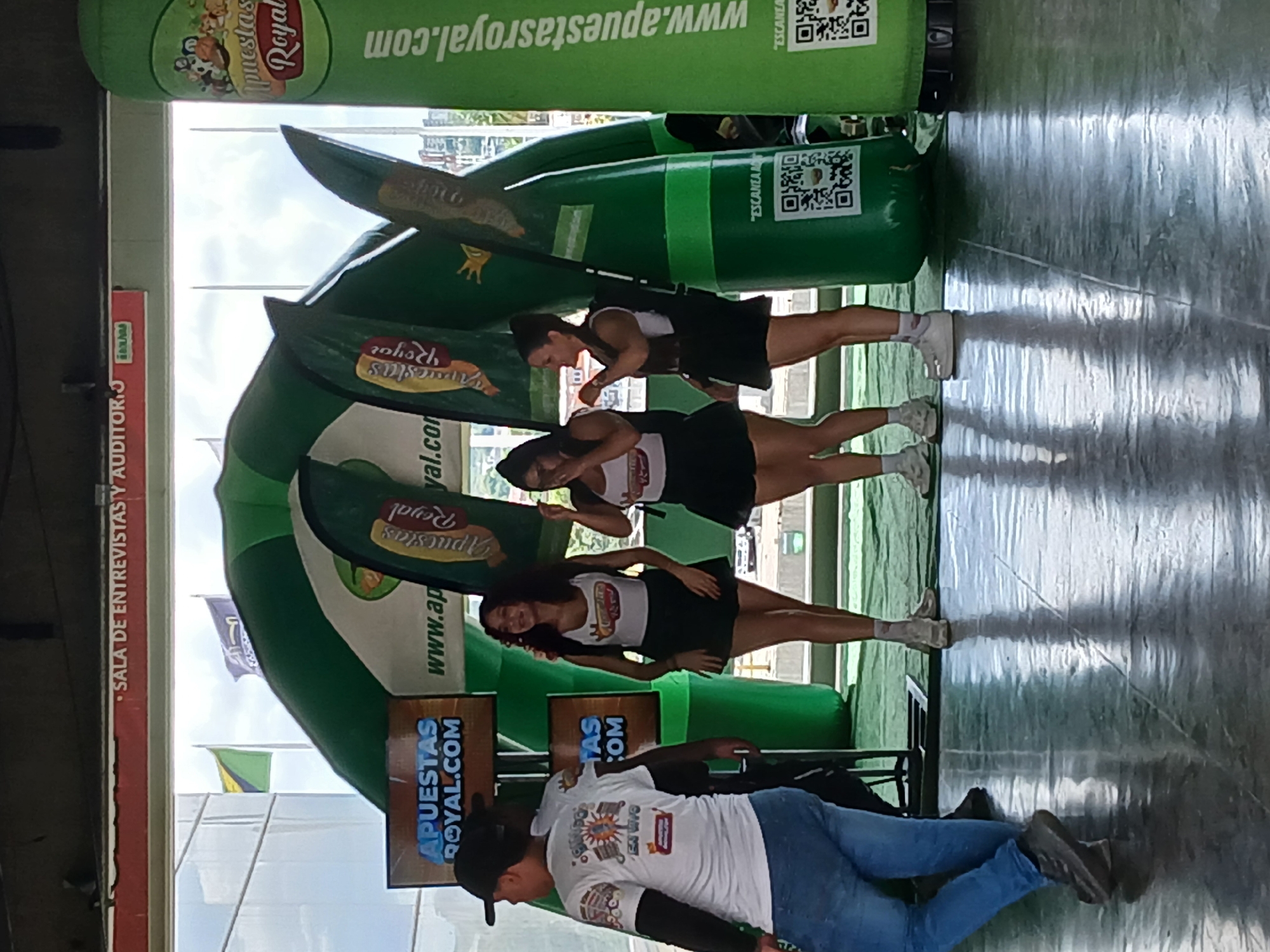The image shows a promotional event in Ciudad Guayana, Venezuela, featuring three women in matching white tops and black skirts posing in front of a large, inflatable green archway. The archway is decorated with the "Apuestas Royal" logo and website address, "www.apuestasroyal.com," also found on banners and signs nearby. A man in a white t-shirt with the same logo is kneeling on the floor. A green inflatable tube stands vertically behind the women, with another website address and QR code. The backdrop shows a building exterior, flags, and the name "SALA DE ENTREVISTAS Y AUDITORIJ." The scene is set on a gray, polished floor, reflecting the surrounding light.