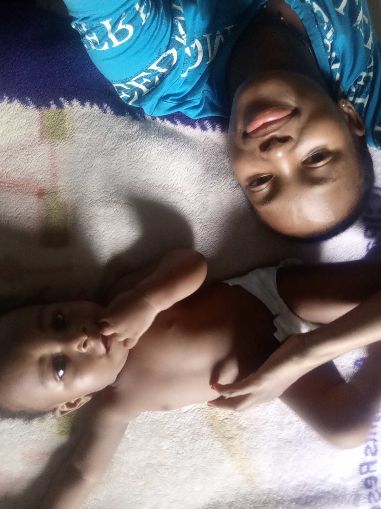 A woman in a blue shirt lies next to a baby on a blanket. The baby is on its back, wearing a diaper. The woman is smiling. The location is Unknown, Unknown.