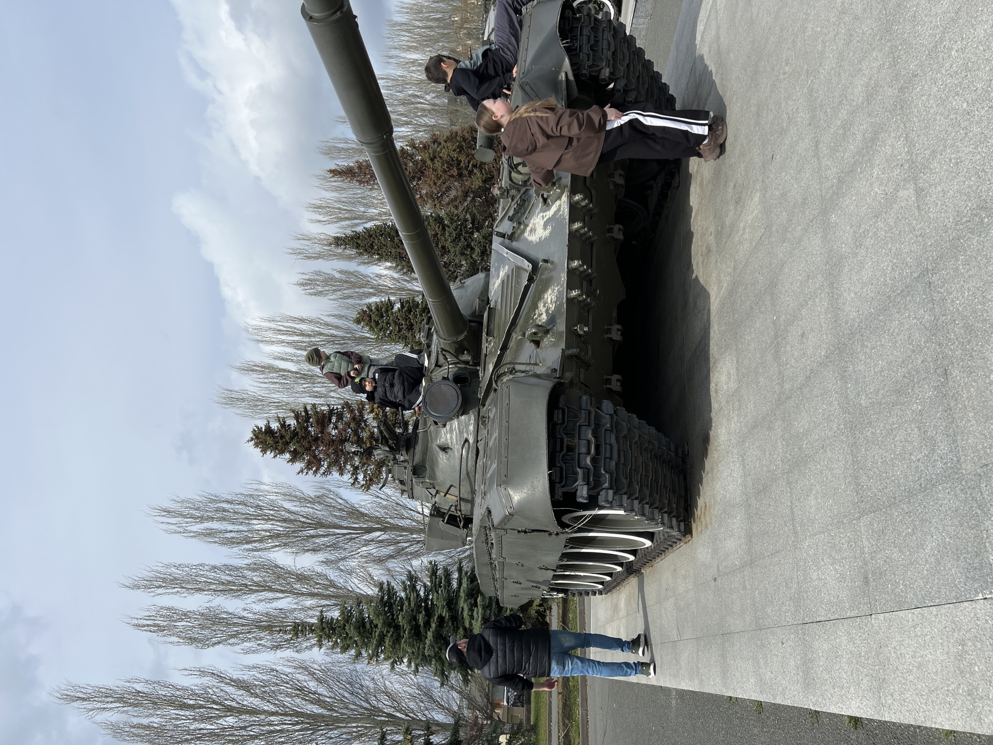 The image displays a large, olive-green military tank positioned on a paved area, likely an outdoor monument or display in Kazan, Russia. The tank, oriented with its long main gun barrel pointing towards the left, features prominent tracks and road wheels.
Several people are interacting with the historical exhibit. Two individuals, appearing to be young people, are on the tank's hull near its turret. One, dressed in a dark jacket, sits atop the tank, while another, in a brown jacket and black track pants with white stripes, stands on the side, observing the tank's features. A third person, partially visible, is also on the tank's surface. On the light grey paved ground next to the tank's tracks, a man in a black puffer jacket, blue jeans, and a baseball cap stands, looking towards the tank.
The background features a mix of tall, bare deciduous trees and dark green evergreen conifers, suggesting a late winter or early spring setting. The sky above is partly cloudy, with visible patches of white clouds against a lighter blue. The activity appears to be a casual visit to the monument, with people exploring and observing the tank. No discernible text is visible on the tank or in the immediate surroundings. The scene takes place during daytime under natural, diffuse lighting.