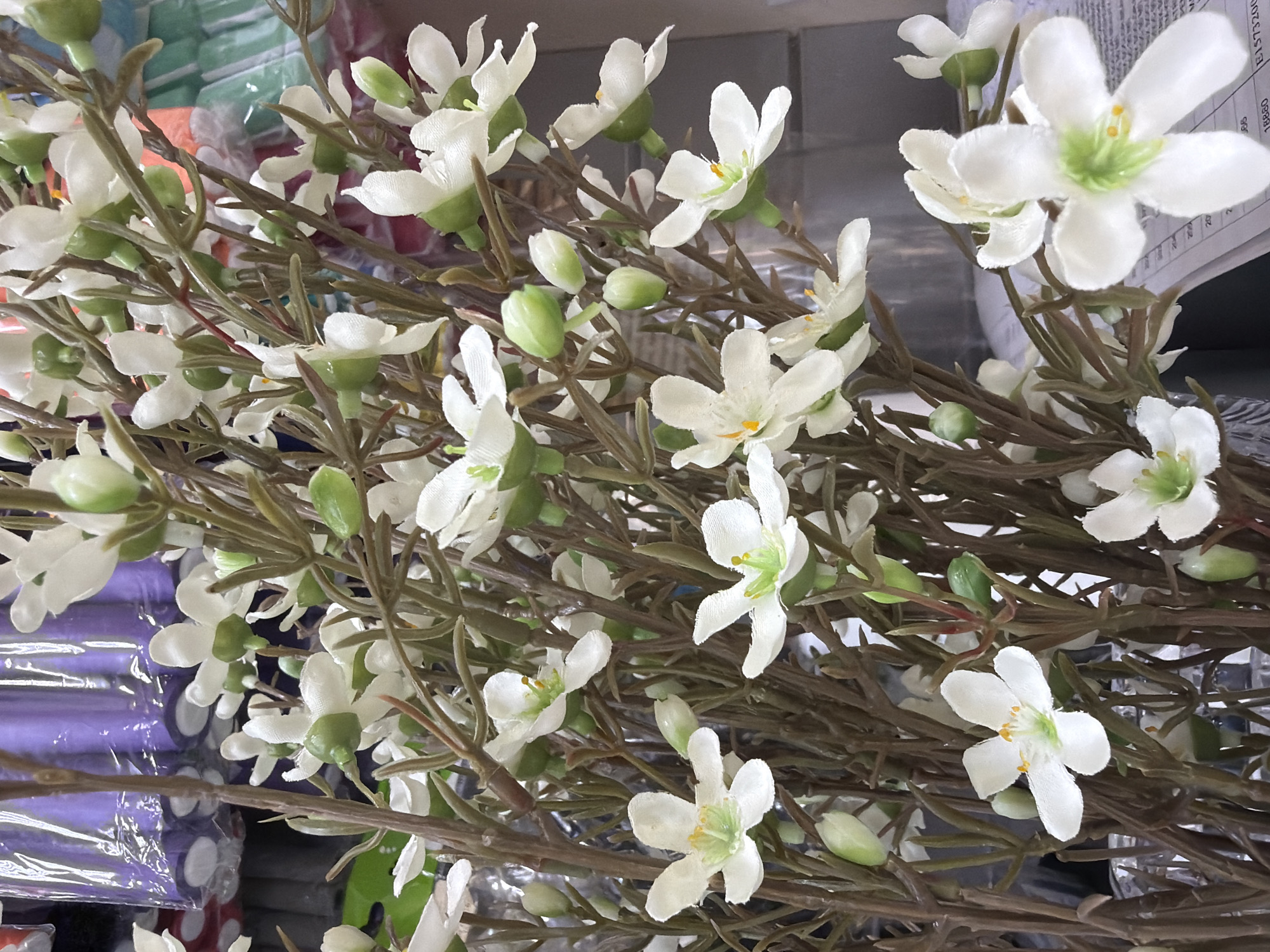 The image depicts a detailed close-up of a dense arrangement of artificial flowers within an indoor commercial setting, located in the city of Sochi, Russia. The primary subjects are numerous white flowers, each featuring distinct white petals surrounding a prominent bright green center, from which delicate yellow or orange stamen-like structures emerge. Many flowers are fully open, while others appear as closed, elongated green buds attached to the stems. The flowers are crafted from a soft, matte fabric, giving them a lifelike texture.
The stems of the flowers are thin, multi-branched, and a uniform dark brown color, resembling dried twigs or bare branches. They are tightly grouped, suggesting a large bouquet or a display of multiple artificial flower bundles.
In the blurred background, particularly in the upper left and central portions, several rolls or bundles of purple material are visible. These items are enclosed in clear plastic packaging, exhibiting a pattern of white polka dots or similar circular motifs. Further to the right in the background, additional blurred items in various colors, including green, light blue, and orange, are discernible, also appearing to be packaged retail goods.
In the bottom right foreground, a portion of a white document or paper is visible. It contains printed text and numbers, specifically fragments like "204," "203," "18880," and "E1573200," suggesting an inventory list, price sheet, or a similar commercial record. The overall lighting is bright and even, consistent with an artificially lit retail environment. No people, explicit actions, or direct interactions are visible within the frame.