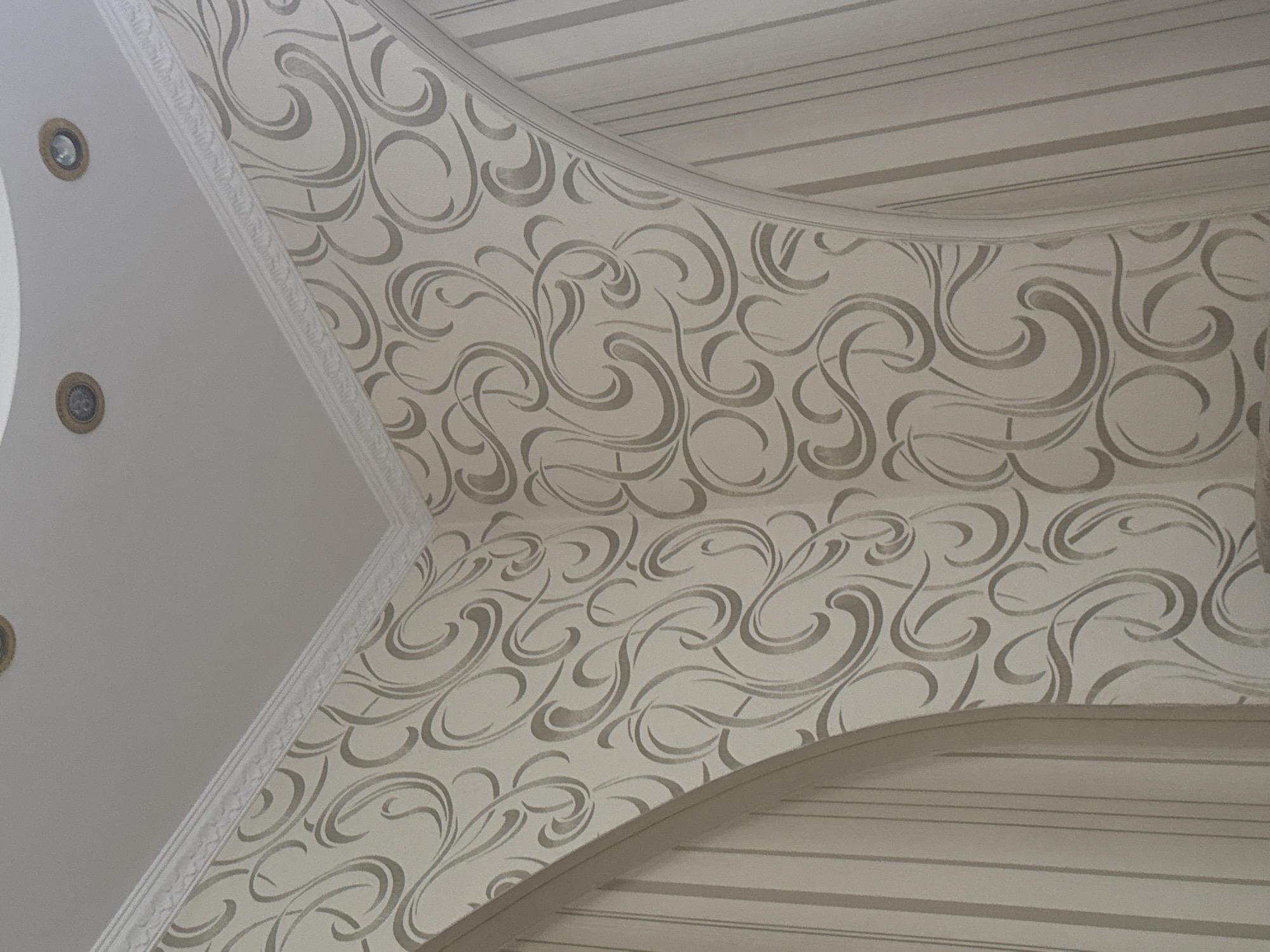 An interior view of a corner where two walls meet and transition into a ceiling. The walls are covered in light-colored wallpaper with a swirling, ornate pattern in a slightly darker shade. Decorative white molding runs along the junction of the walls and the ceiling. The ceiling is white and features two recessed circular light fixtures. On either side of the central patterned wall, there are curved archways leading to areas covered in vertically striped wallpaper. The overall impression is of an interior architectural detail with decorative finishes.