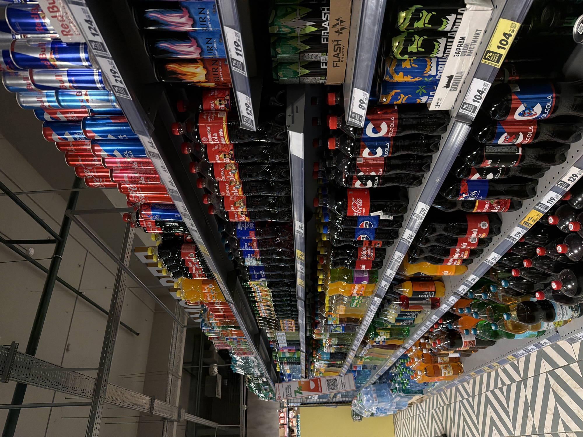 The image shows a supermarket shelf stocked with various beverages in Izhevsk, Russia. Canned and bottled drinks, including energy drinks (Red Bull, Burn, Flash), colas (Coca-Cola, Pepsi, Dobry Cola, Ol Cola), and other soft drinks, are arranged on multiple shelves with price tags displayed. The shelving system is made of metal, and the floor has a patterned tile design. No people are visible in the frame.
