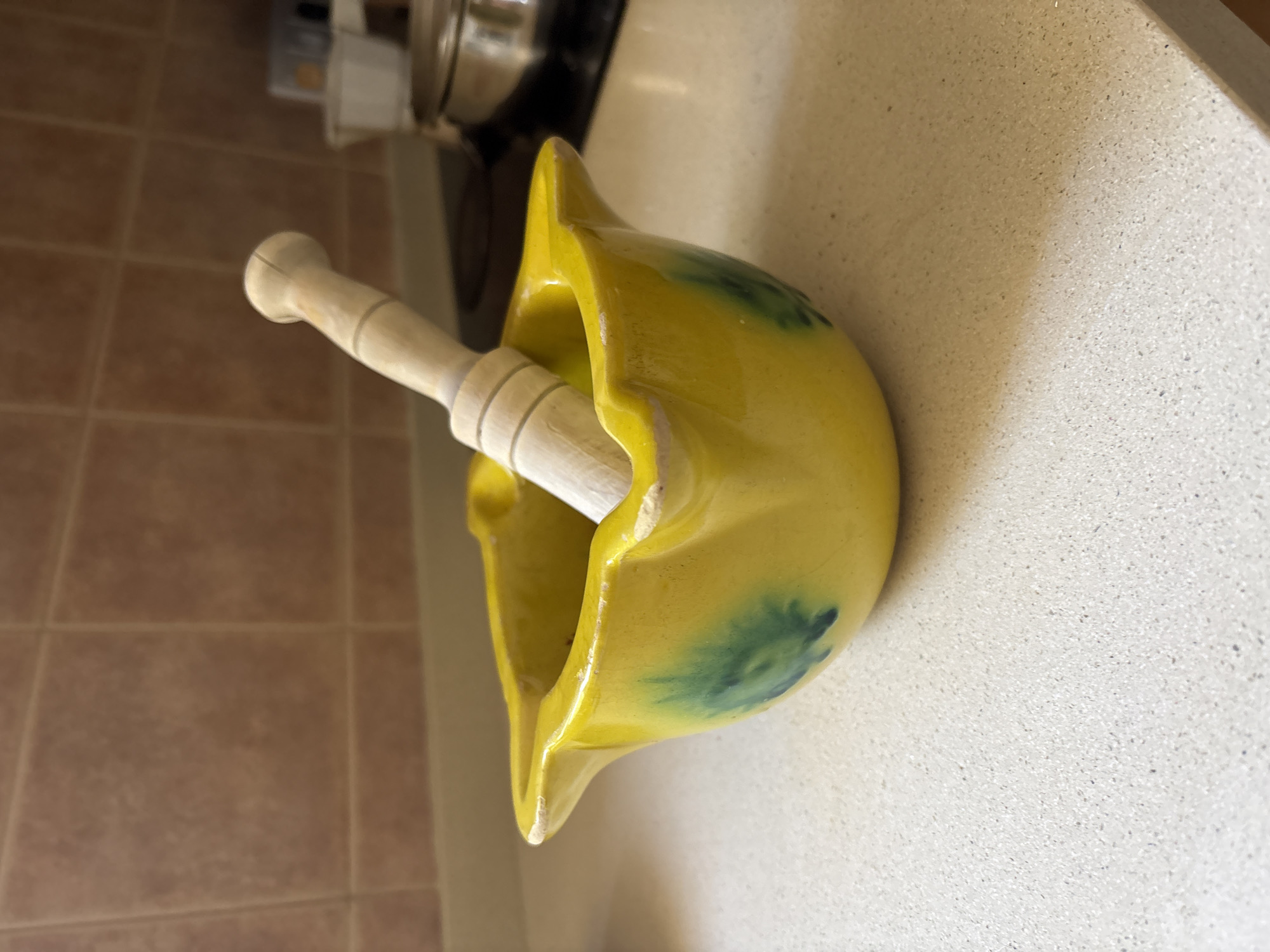The image shows a yellow ceramic mortar with a wooden pestle inside. The mortar has a flared rim and some green discoloration on its surface. The pestle is cylindrical with a rounded end. The mortar and pestle are sitting on a light-colored countertop, possibly in a kitchen. The background includes brown tile flooring and a glimpse of a stainless steel pot or pan. While it's difficult to definitively confirm, the location is likely a residential setting within Urbanización Encinares De Alcolea, Spain, based on the provided information.