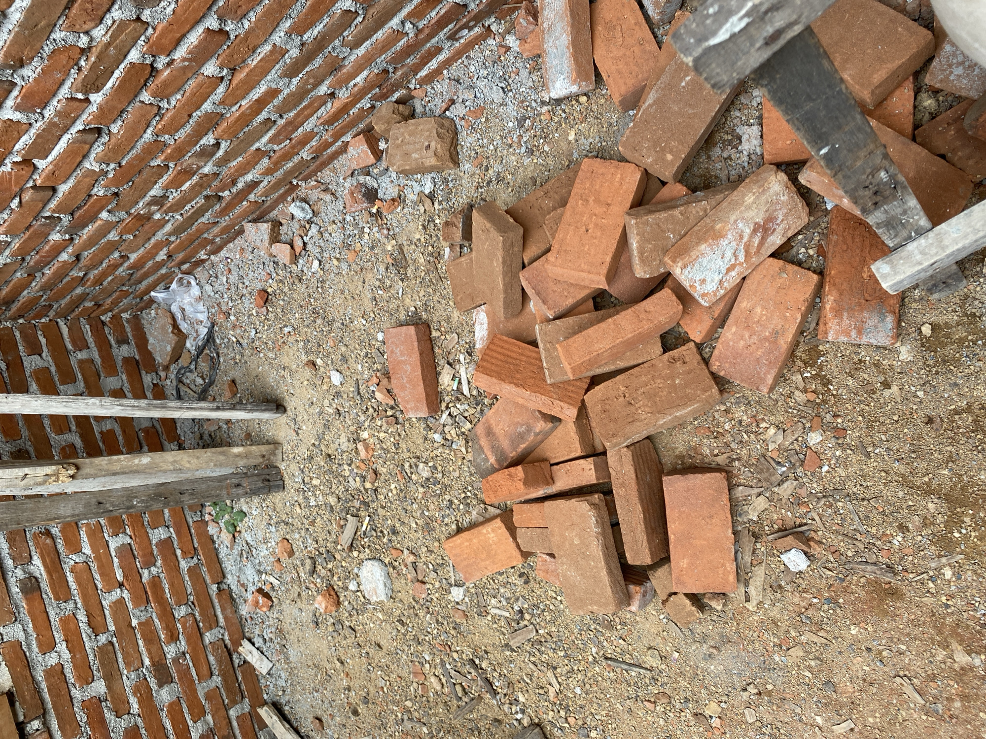 The image shows a construction site in Banda Aceh, Indonesia. The scene is an unfinished room with brick walls. A pile of red bricks lies on the sandy and gravelly floor. Wooden planks are propped against the walls and scattered around. There are no people visible in the image. The environment appears to be outdoors or an open construction area.