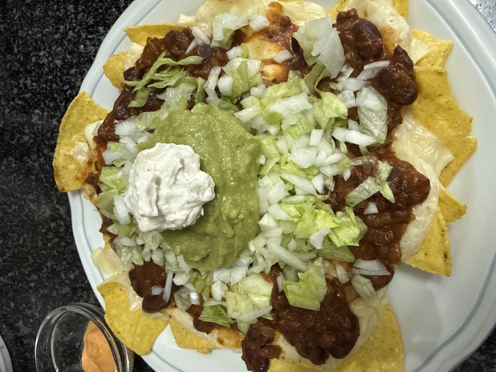 A plate of nachos sits on a counter. The nachos are topped with cheese, chili, guacamole, sour cream, shredded lettuce, and chopped onions. A small bowl containing orange sauce is visible near the plate. The counter is a dark color.