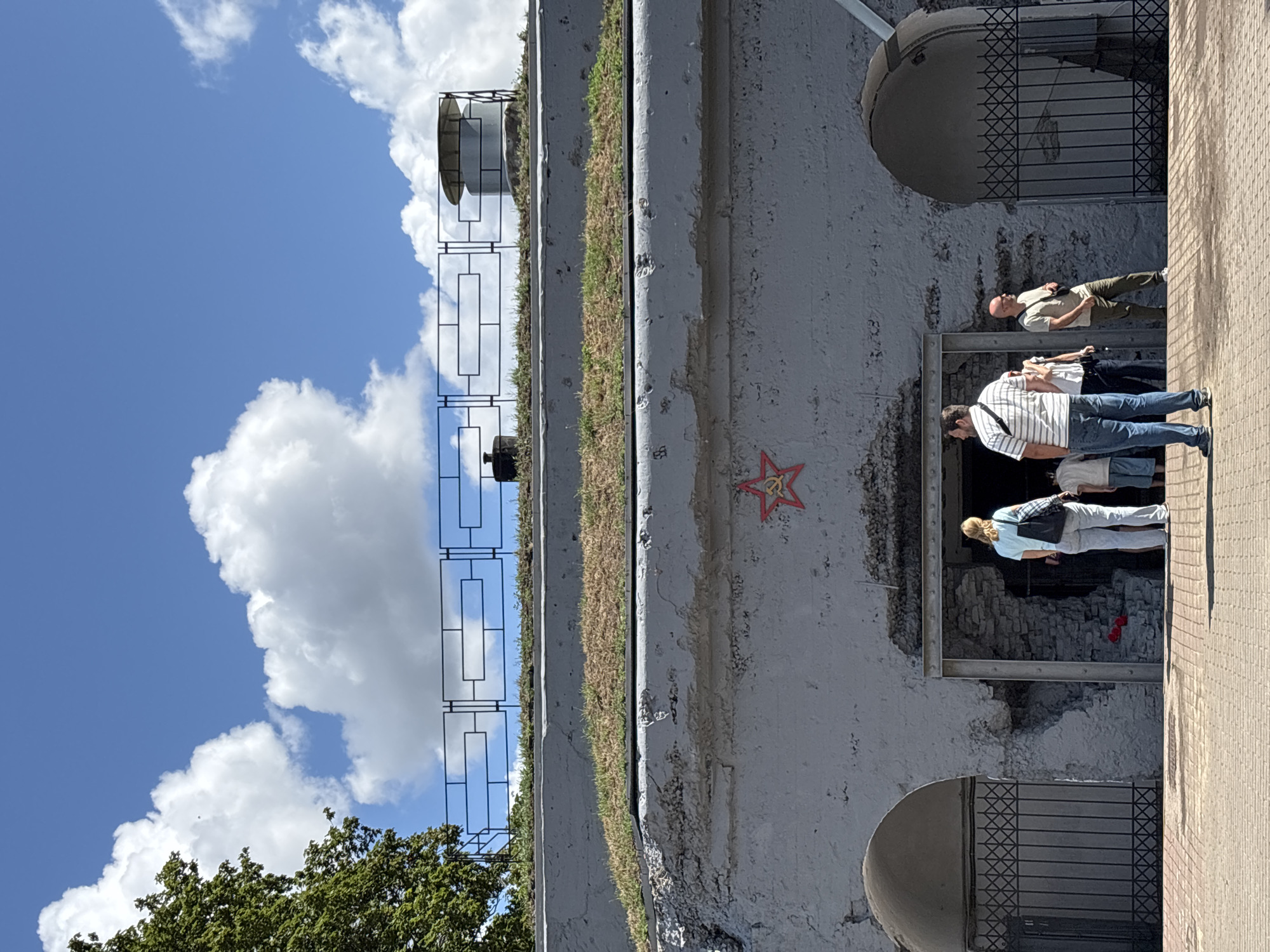 The image captures the entrance to the Brest Fortress in Brest, Belarus. In the foreground, a brick-paved pathway leads to the fortified structure. Several individuals are visible, some standing and others walking, near the entrance. The fortress itself is constructed of grey stone and concrete, with arched openings and visible signs of weathering. A red star emblem, a symbol associated with the Soviet Union, is affixed to the upper portion of the facade. Above the fortress, a bright blue sky is dotted with fluffy white clouds. A metal railing runs along the top of the fortress wall, and a cylindrical structure, possibly a vent or a lookout post, is also visible. Lush green grass grows on the sloped roof of the fortress.