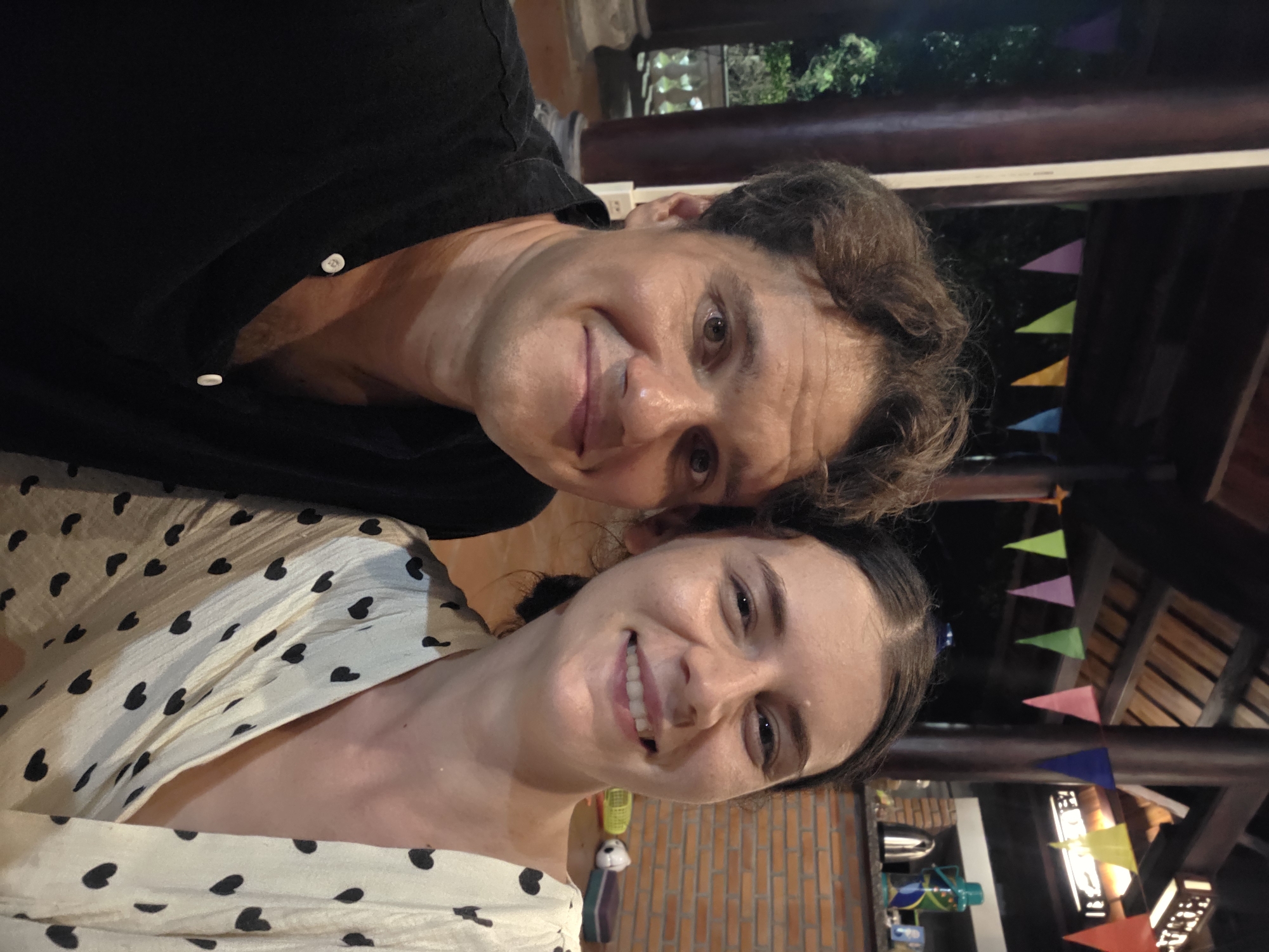Two people are posing for a selfie indoors, with a man on top wearing a black shirt and a woman below wearing a white shirt with black heart patterns. Behind them are decorations of colorful triangular flags hanging from wooden beams. In the background, there is a brick wall and other items suggesting a casual dining or gathering space. Based on the location, this could be a restaurant or event space in Mũi Né, Vietnam, given the presence of common architectural elements found in the region.