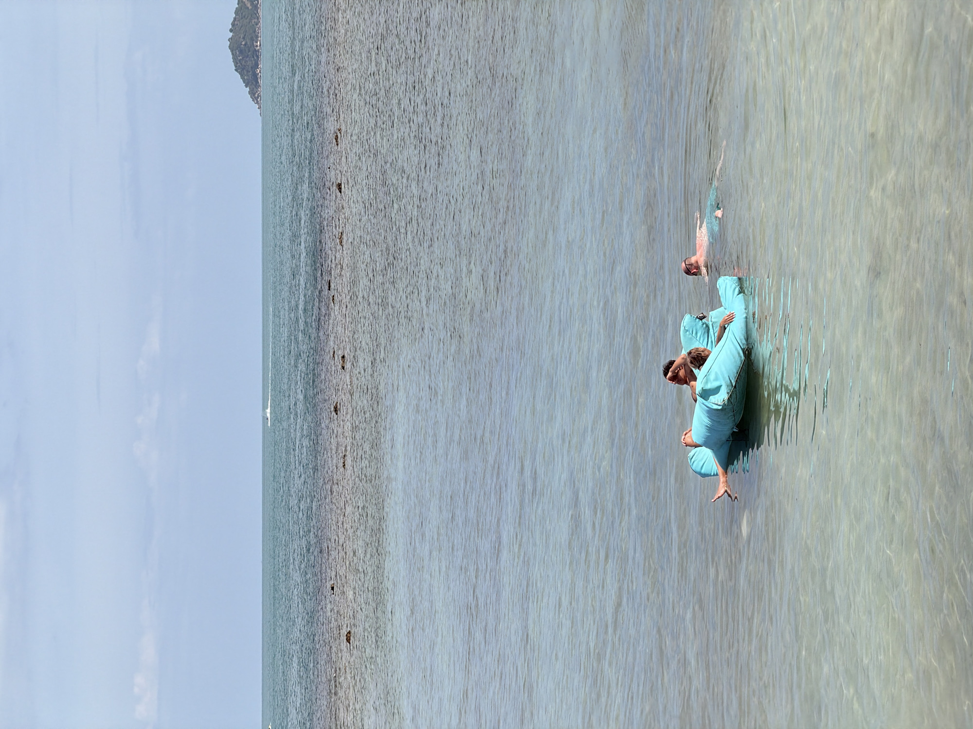 In the clear, shallow waters off the coast of Ko Samui District, Thailand, four individuals are seen enjoying a leisurely day. A bright turquoise inflatable raft, shaped like a crescent moon, floats on the surface. Two people are lounging on the raft, one with their legs extended and the other appearing to be resting their head on a pillow. A third person, a man wearing swimming trunks, is swimming nearby, with his body partially submerged. A fourth individual, also swimming, is in the background. The calm, translucent water reveals the sandy bottom below. In the distance, a small, tree-covered island rises from the horizon, and a white boat is visible on the sea. The sky above is a pale blue, with scattered white clouds. The overall scene depicts a serene and relaxing day at the beach.